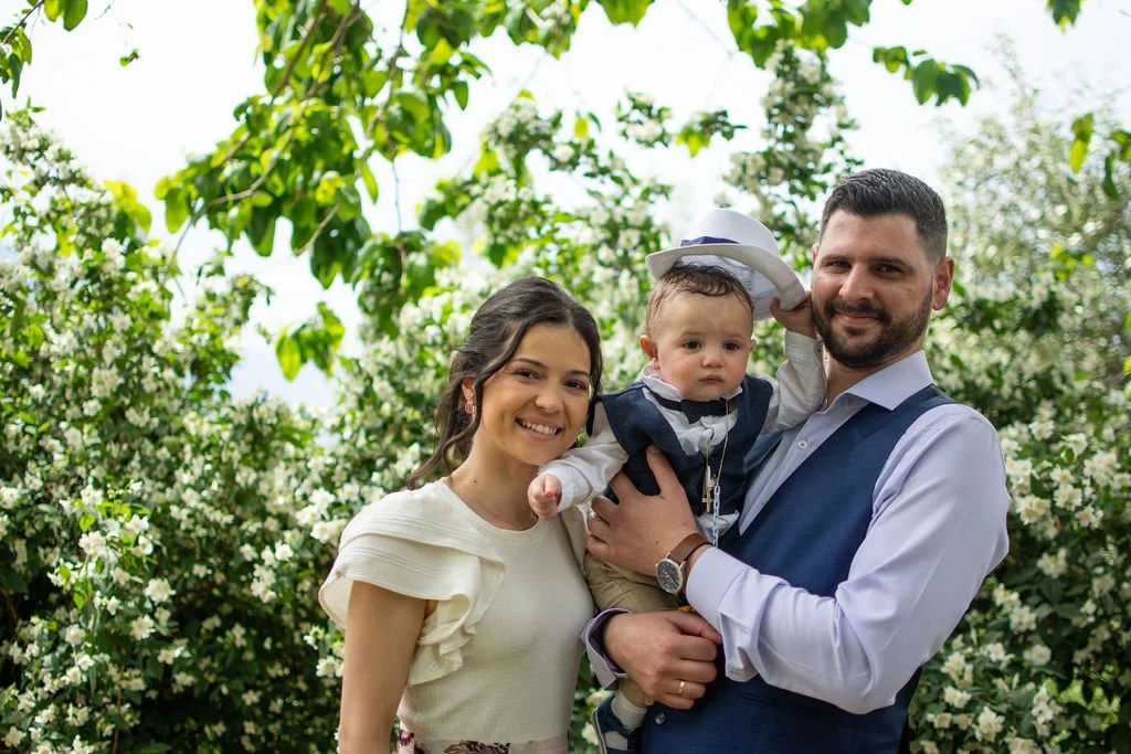 A happy family of three posing outdoors in front of blooming trees. The mother joyful, the father holding a young child in formal attire. The child has a small hat held by the father.