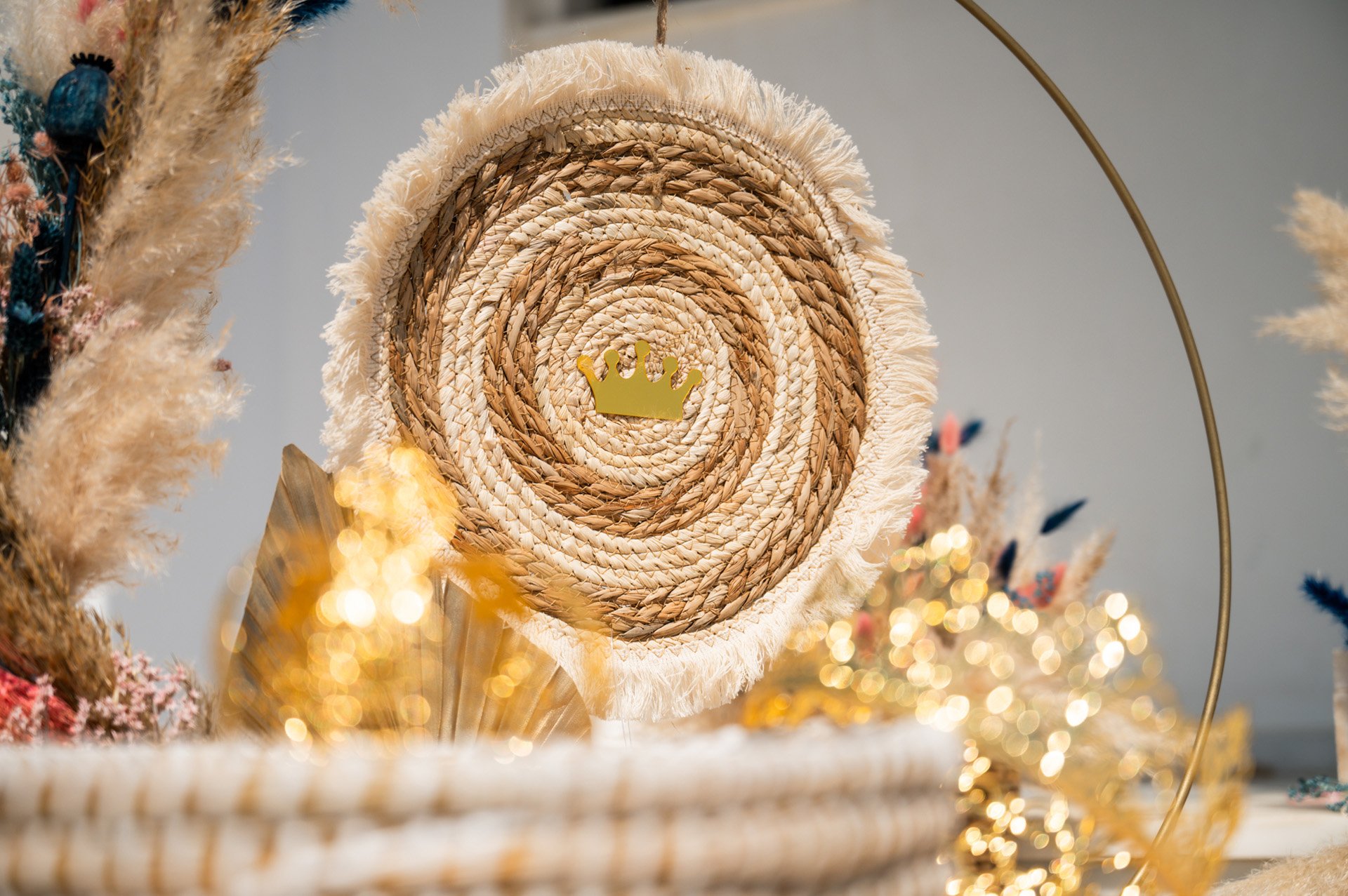 A decorative woven wall hanging with a gold crown in the center, surrounded by ornamental dried flowers and golden accents.