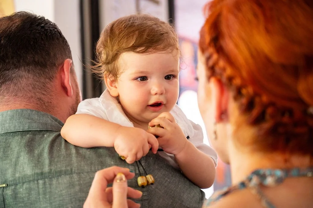 A young child with curly hair sitting on an adult's shoulder, looking at a woman with red hair, in an indoor setting.