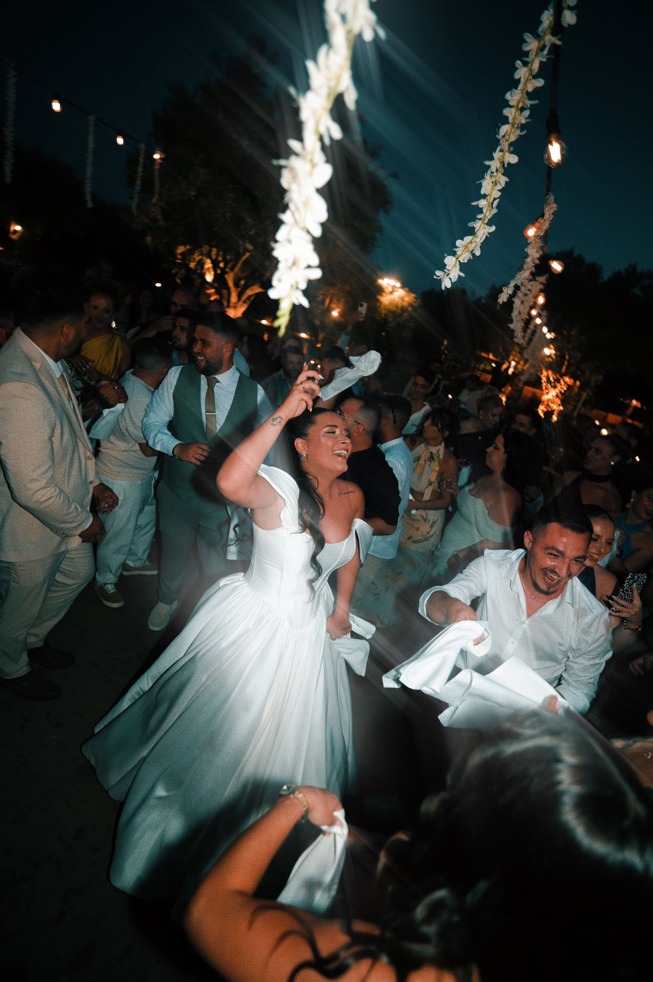 A bride and groom dancing at an outdoor wedding reception at night, with guests surrounding them and hanging string lights and flowers above.