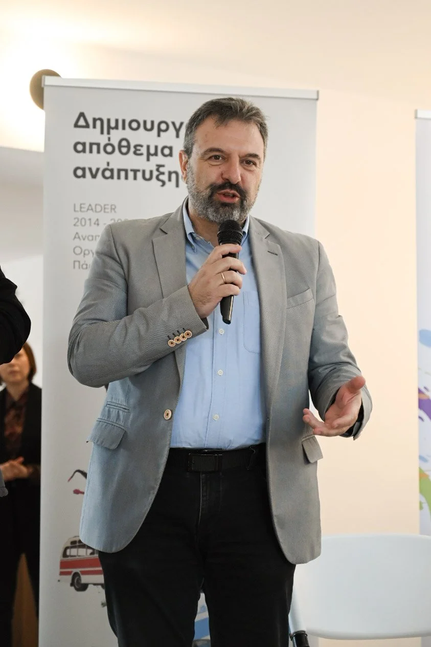 A man in a grey blazer and blue shirt speaking into a microphone at an event, standing in front of a poster with Greek text.