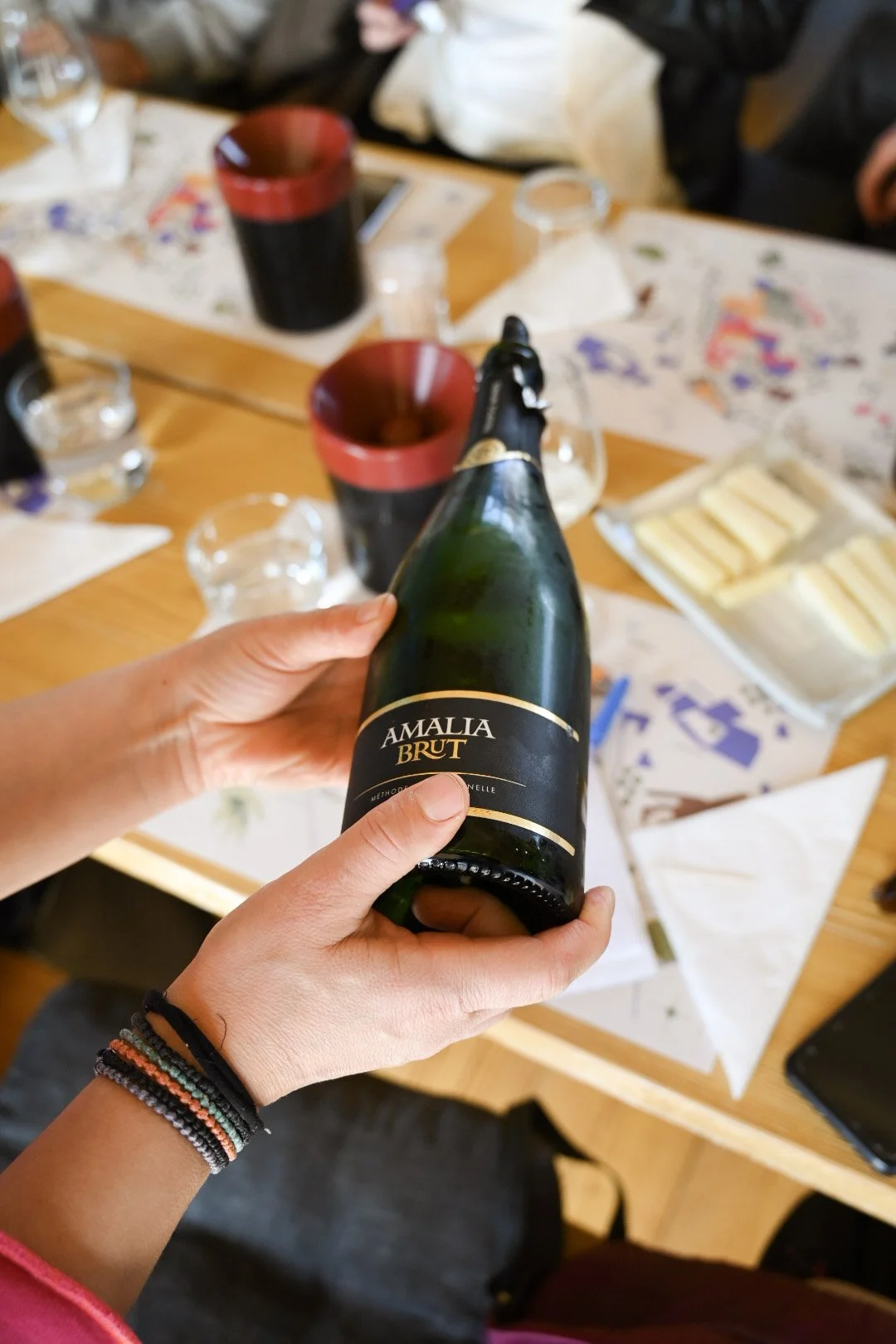 Person holding a bottle of Amalia Brut sparkling wine over a table set with glasses, napkins, and a tray of cheese.