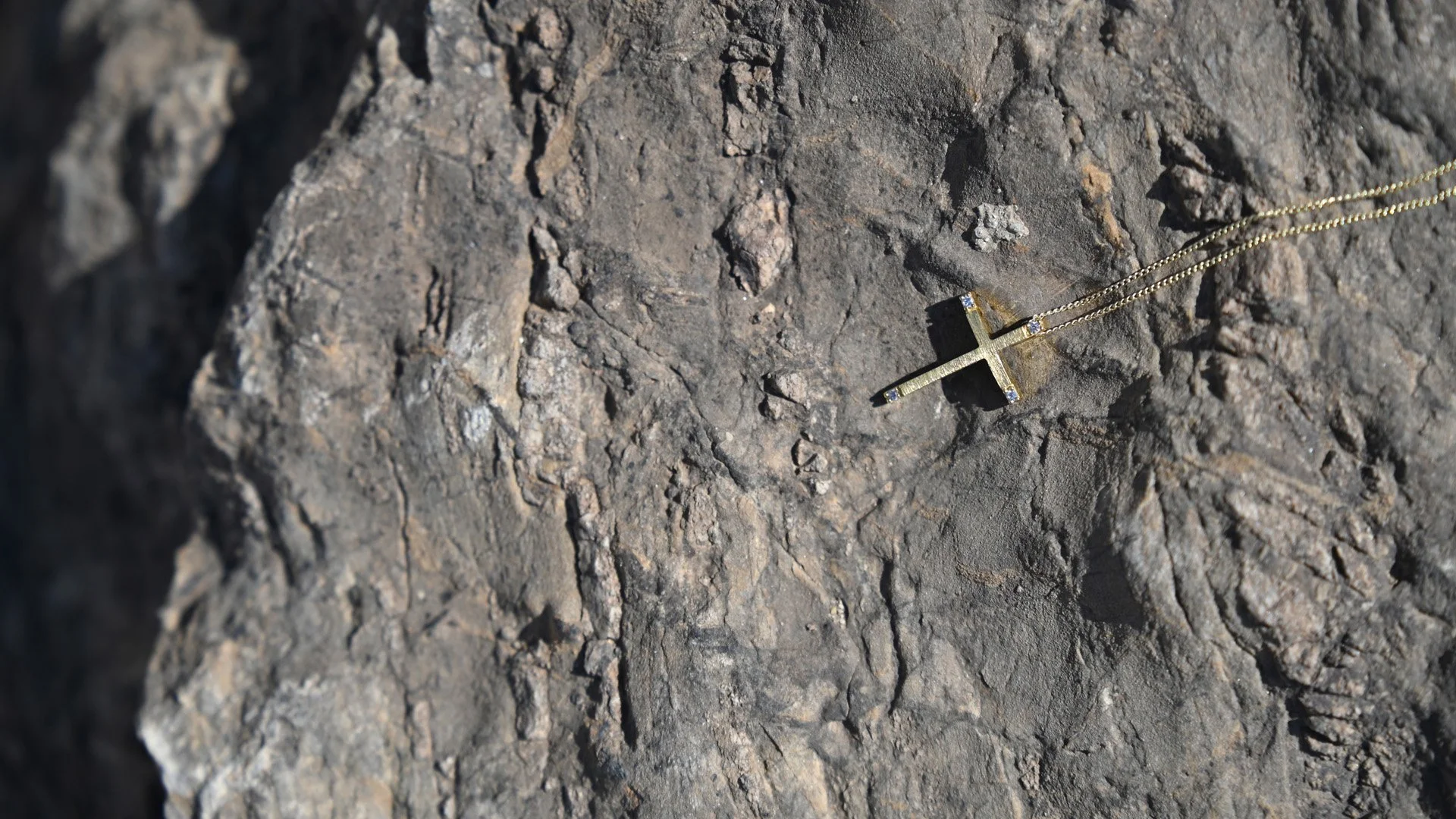 Gold necklace with cross pendant lying on dark textured rock surface.