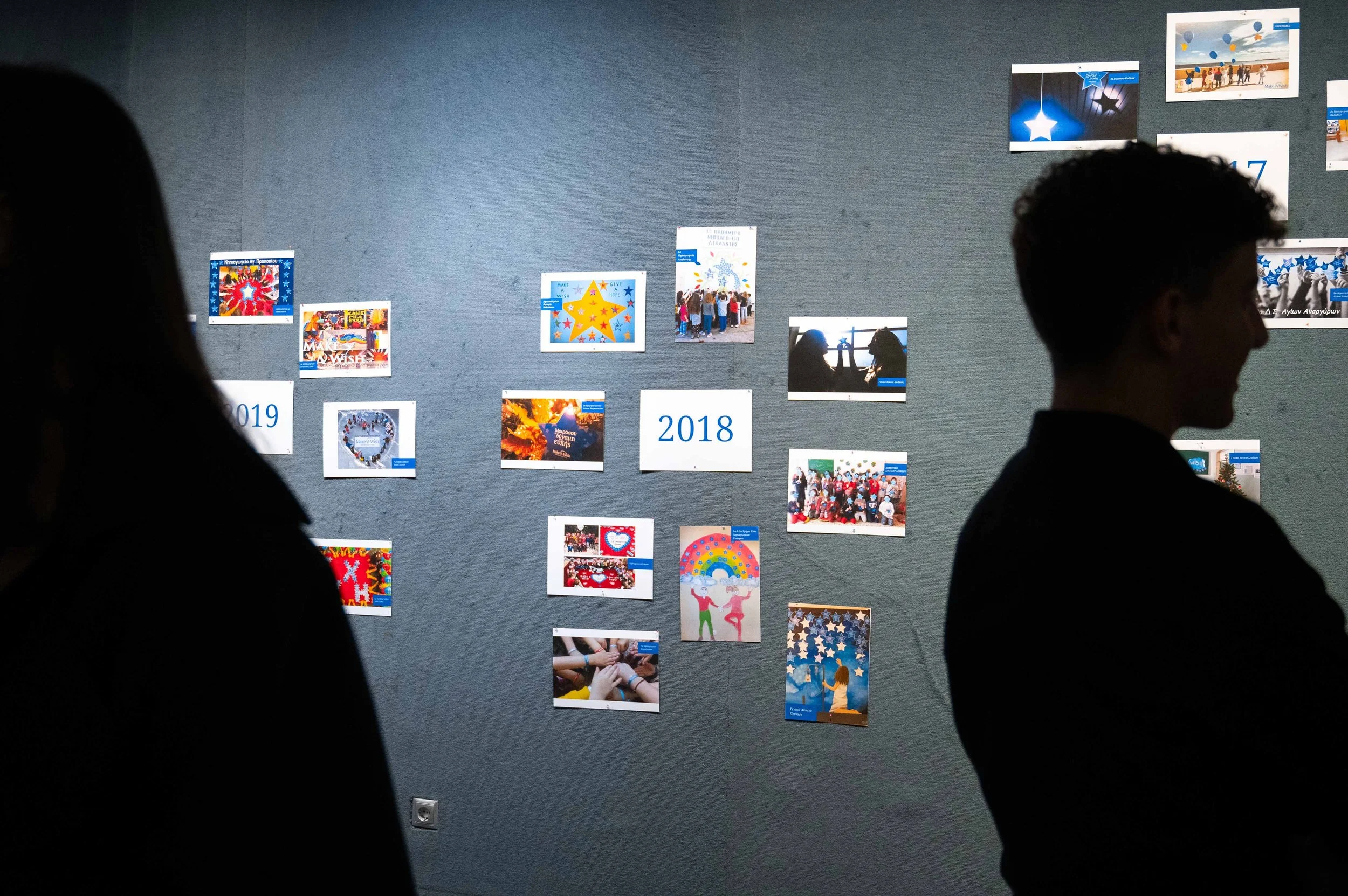 People standing in front of a wall with various photos and posters, some labeled with the years 2018 and 2019.