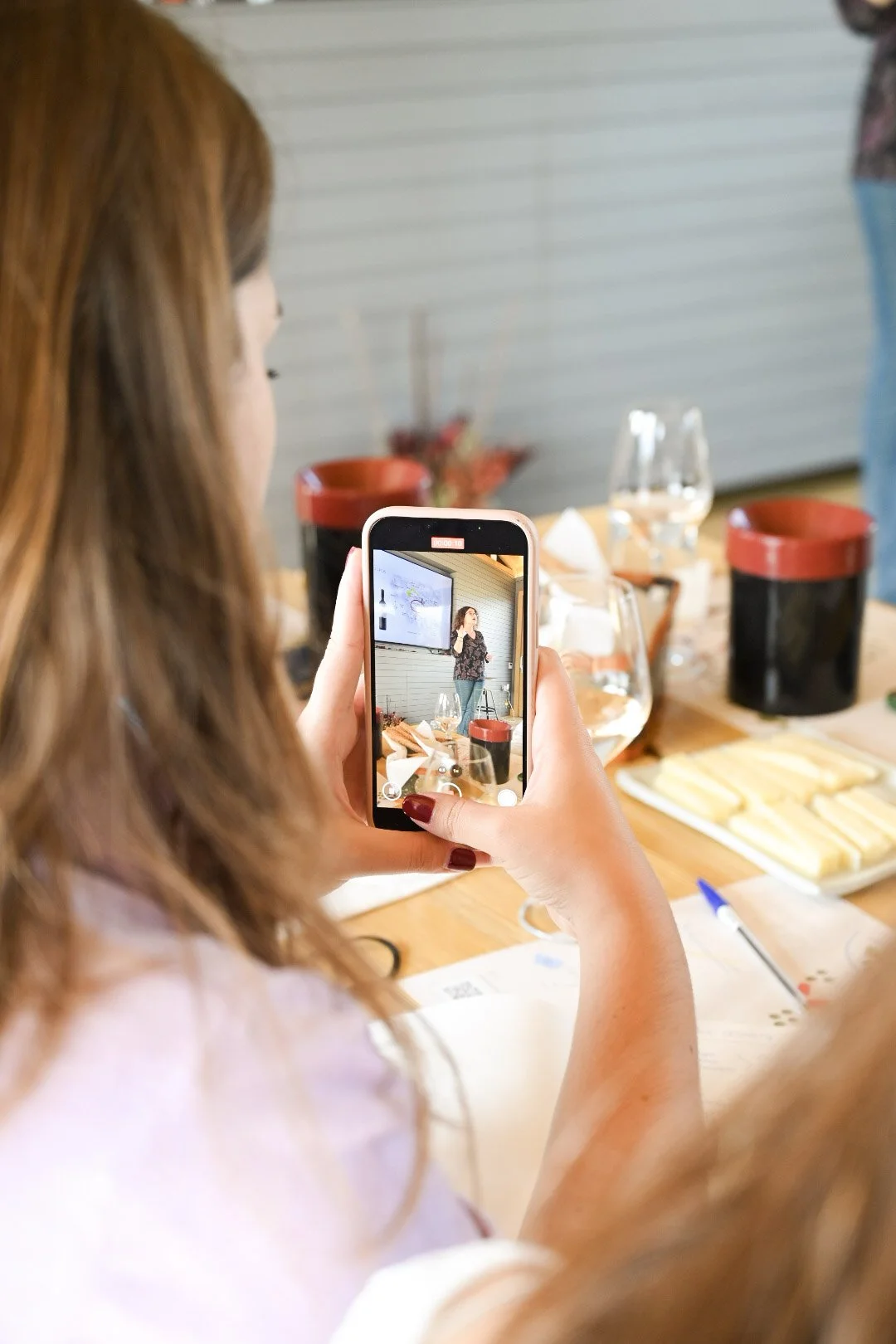 A woman with brown hair taking a photo with her smartphone of another woman speaking in front of a screen in a meeting room.