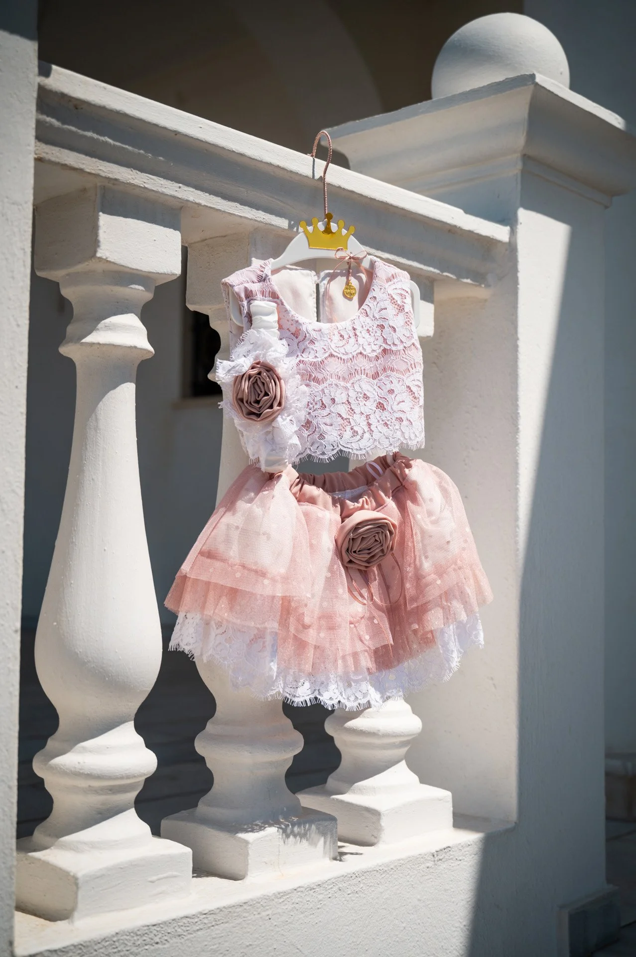 A children's dress hanging on a white railing outside. The outfit includes a pink lace top and a pink tutu with lace and fabric flowers, decorated with ribbon bows.