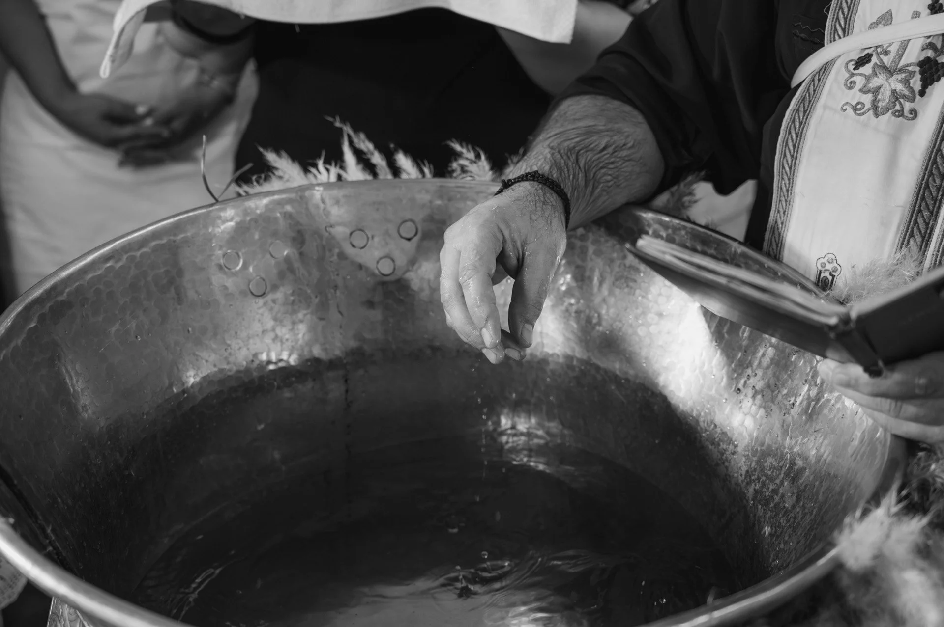 A person’s hand dips into a large metal basin filled with liquid, while another person observes and holds a pamphlet.