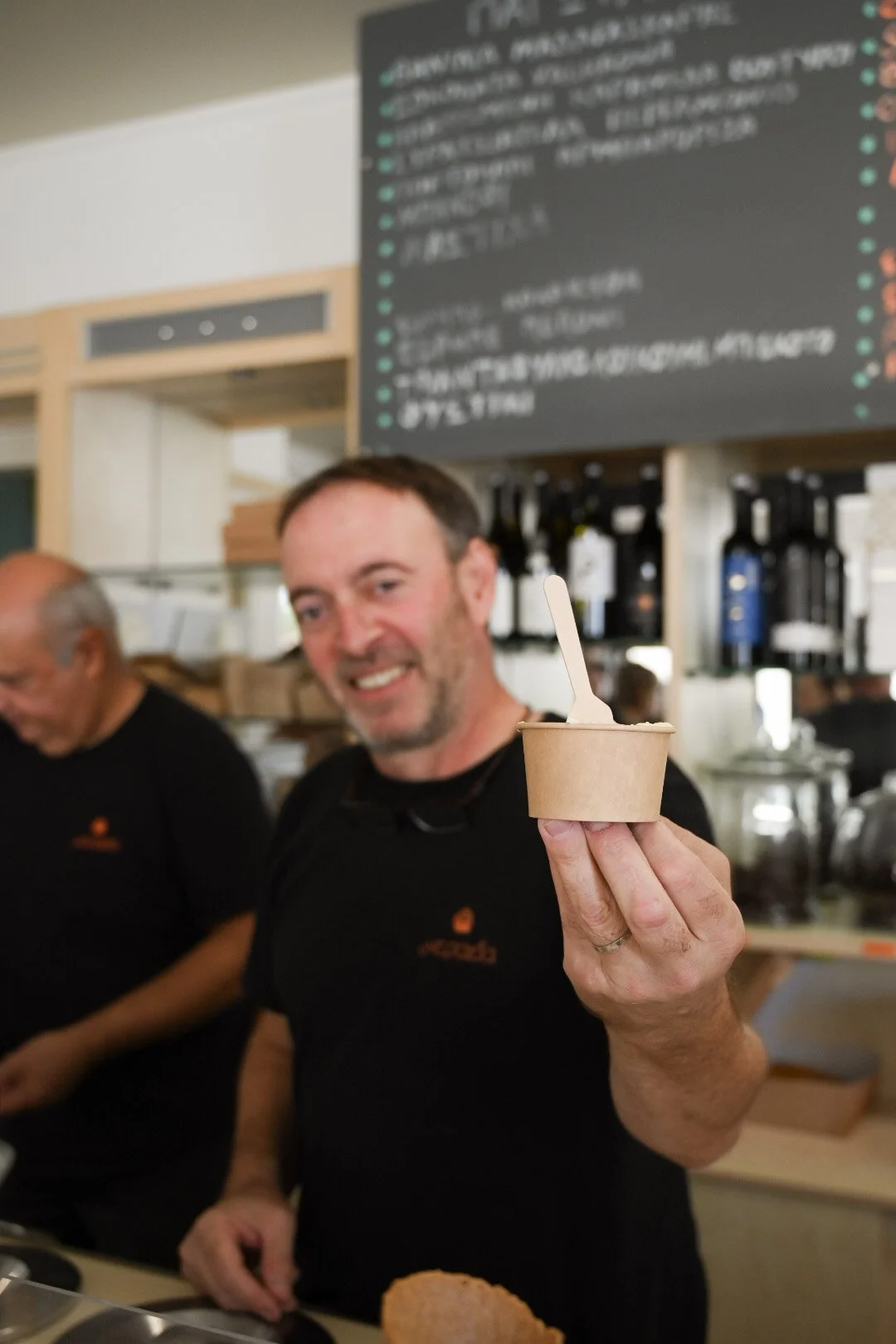 Smiling man holding a small cup of ice cream with a white spoon inside at a cafe or restaurant.