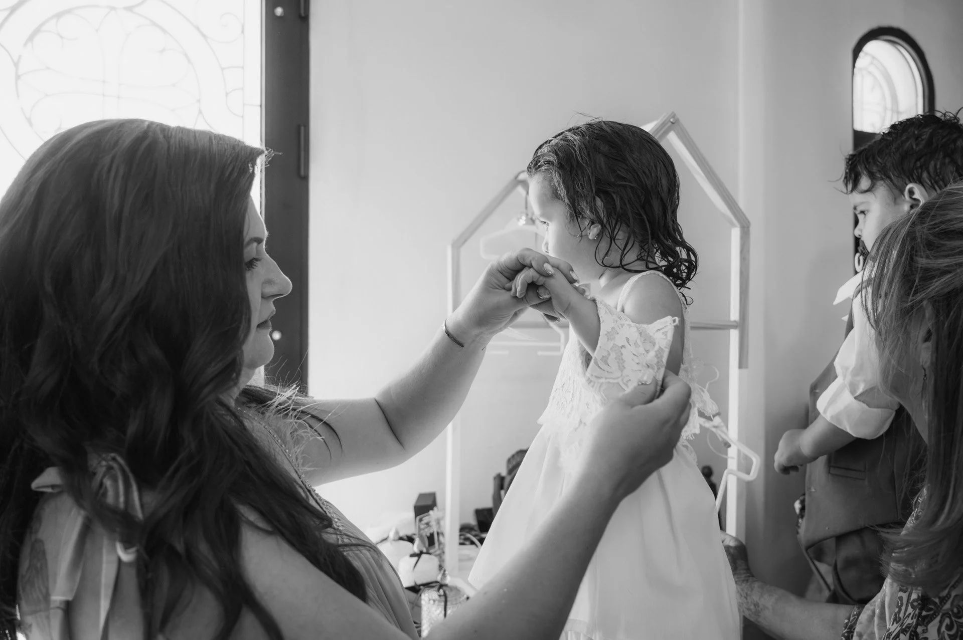A woman helps a young girl in a white dress prepare for a special occasion, possibly a wedding, with other people assisting nearby.