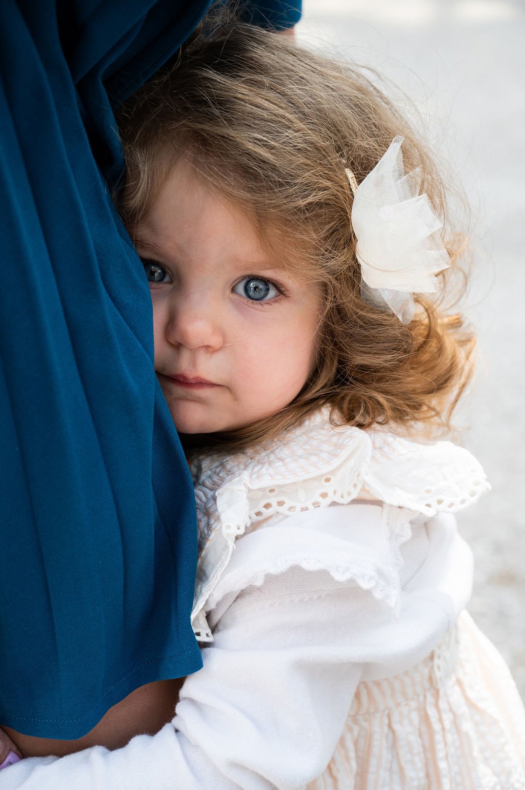 A young girl with curly light brown hair and blue eyes, wearing a white dress with lace details and a white bow in her hair, resting her head against an adult who is partially visible, with a blue sleeve. She is looking directly at the camera.