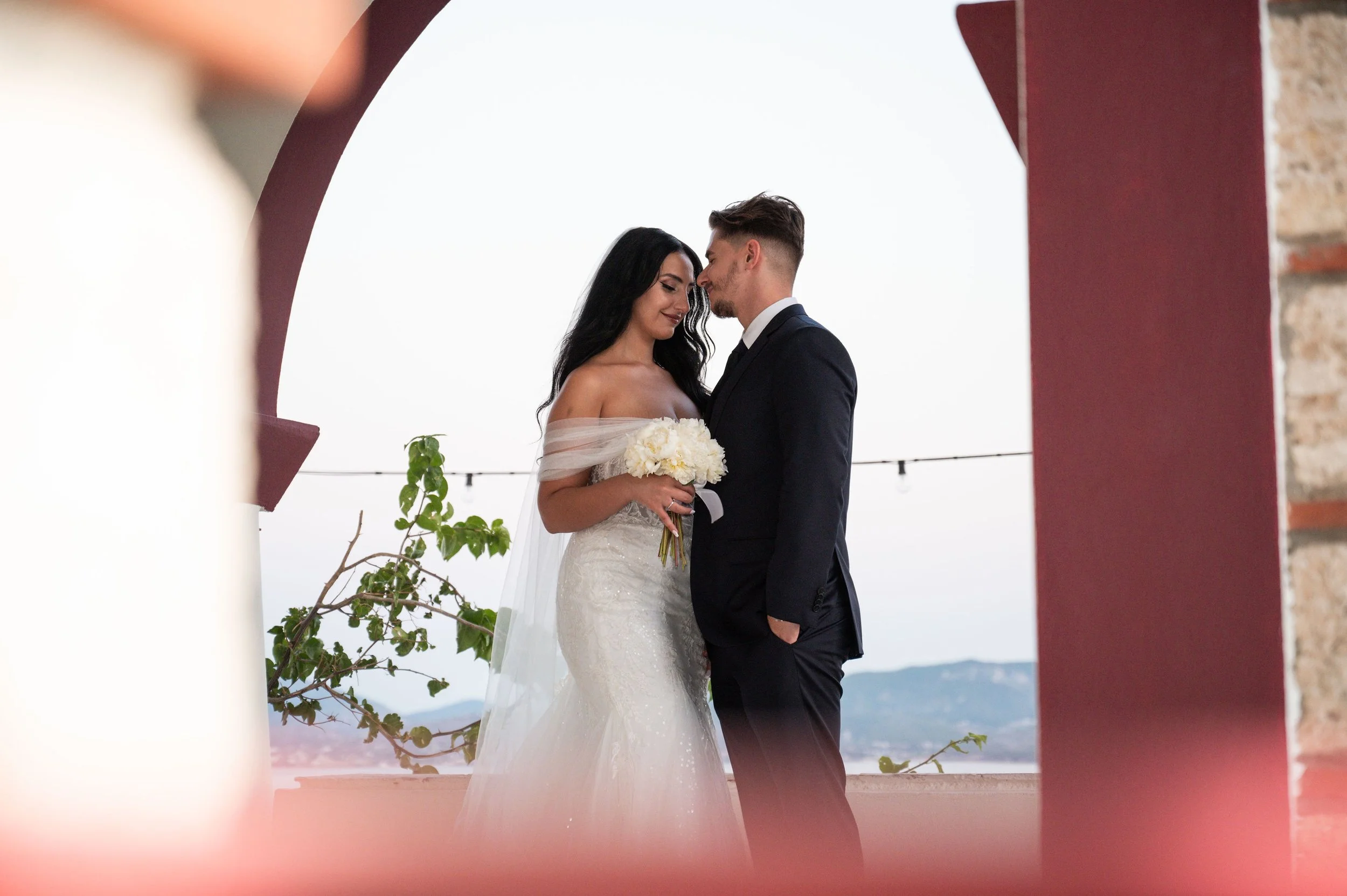 A bride and groom standing close together outdoors during their wedding, with the bride holding a bouquet of white flowers and the groom in a dark suit, both smiling softly, framed by an archway.