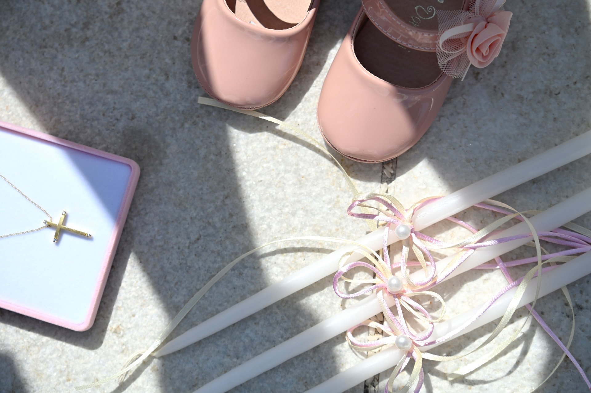 Pink baby shoes with decorative flowers and tulle, a necklace with a cross cross pendant in a pink box, and white candles with pink and cream ribbons and pearl embellishments on a gray surface.