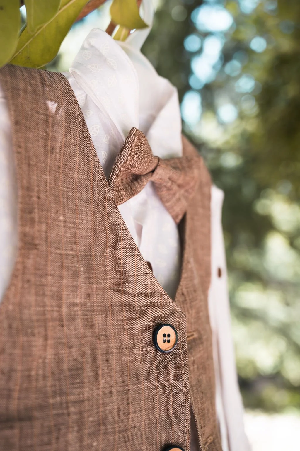 Close-up of a mannequin dressed in a white shirt and a brown fabric vest with matching bow tie, outdoor setting with blurred trees and sunlight in the background.