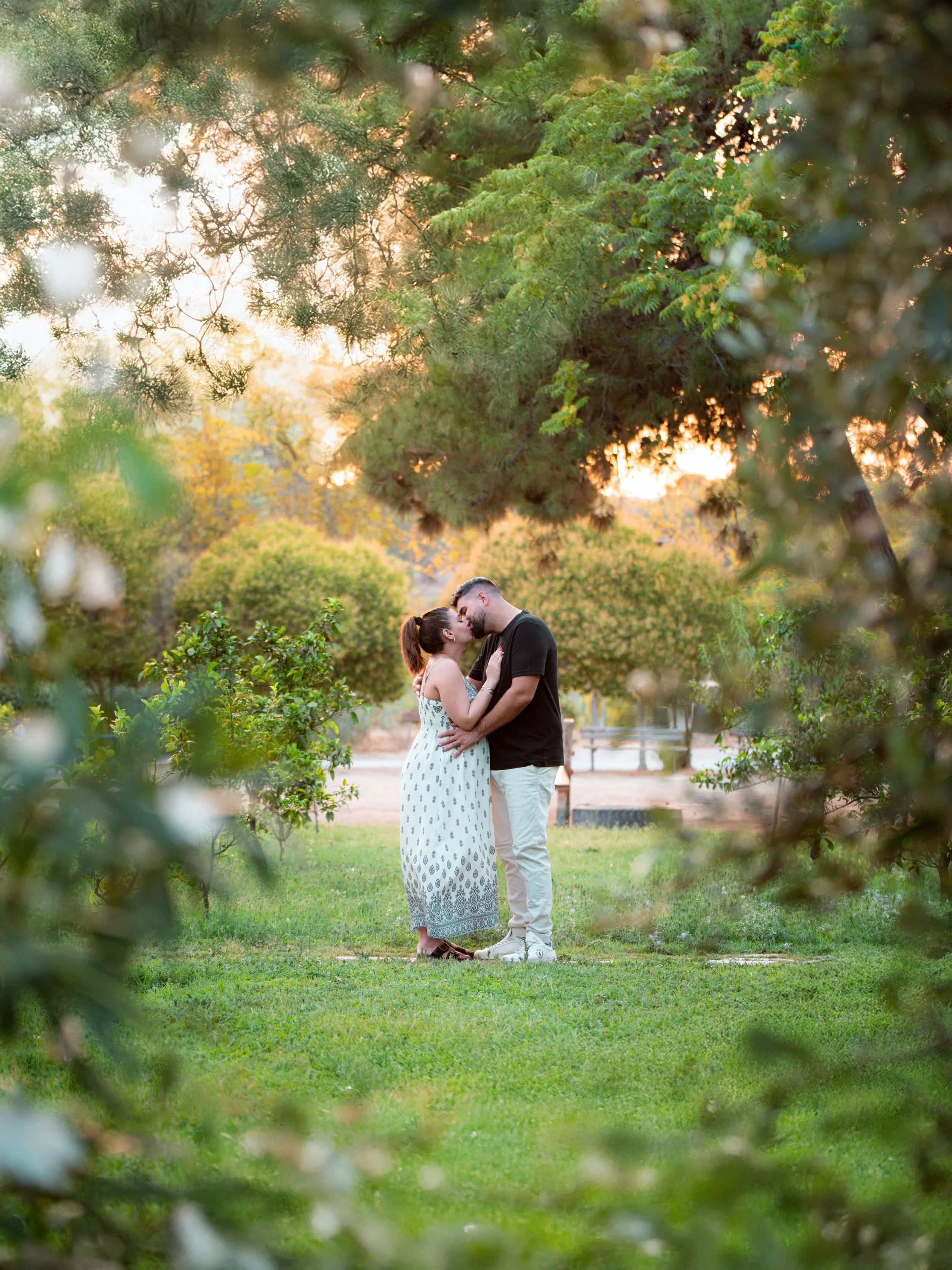A couple sharing a kiss in a park during sunset, framed by green trees and bushes.