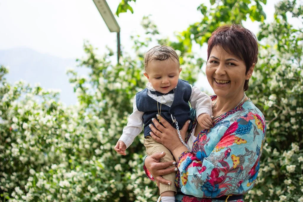 A smiling woman with short dark hair holding a young child outdoors, with greenery and white flowers in the background.