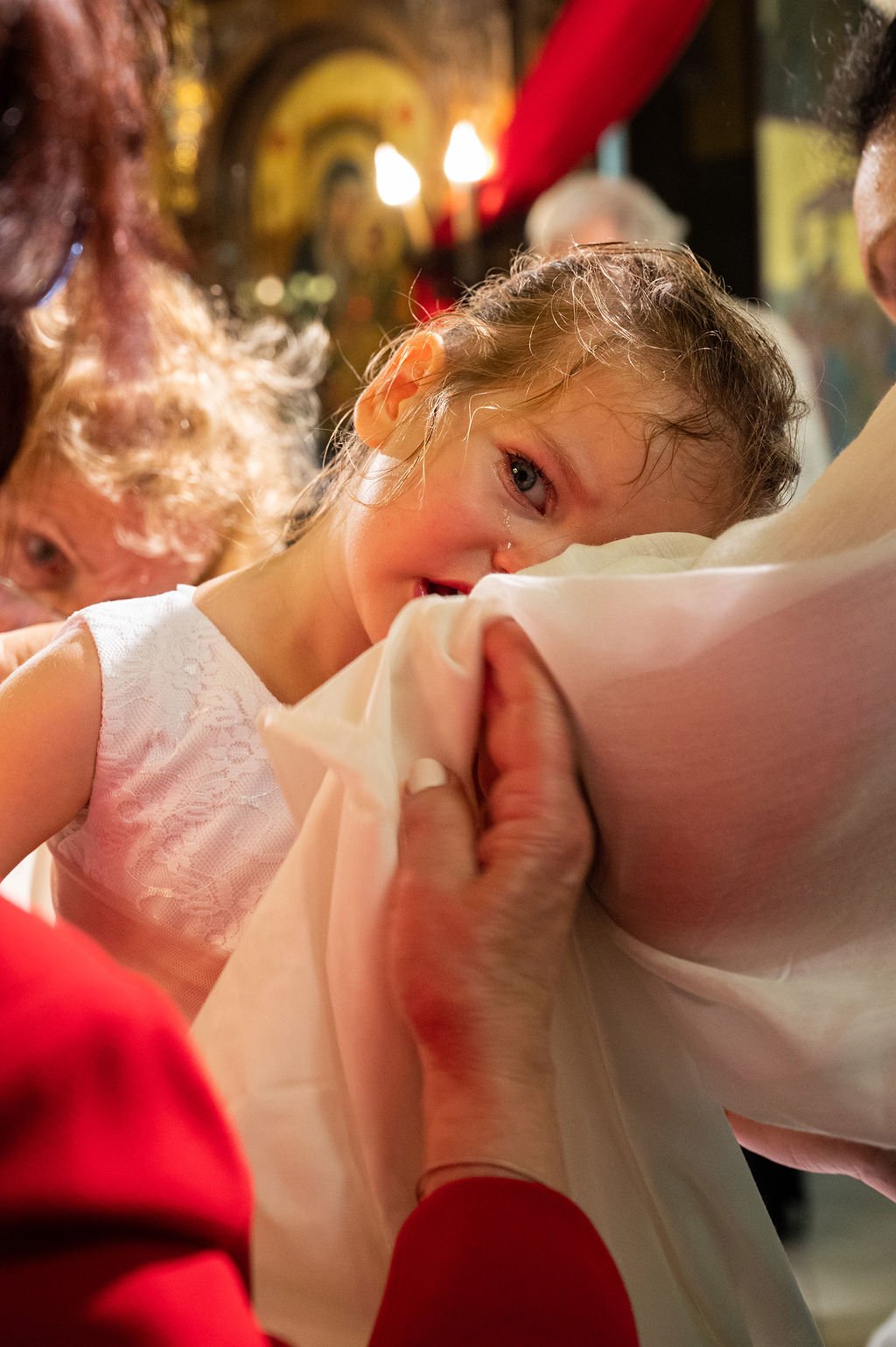 A young girl in a white lace dress happily receiving a christening blessing from a priest, with an elderly woman in the background and warm church lighting.