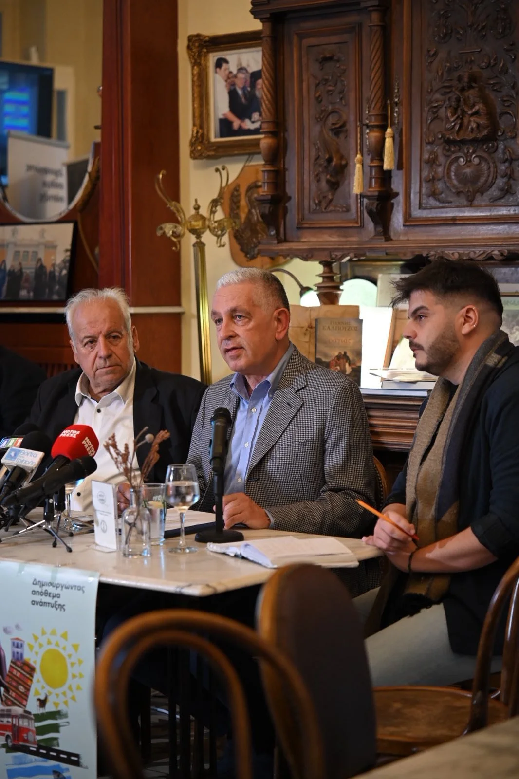 Three men seated at a table during a press conference, with microphones in front of them, in a room decorated with wooden furniture and framed photographs on the wall.