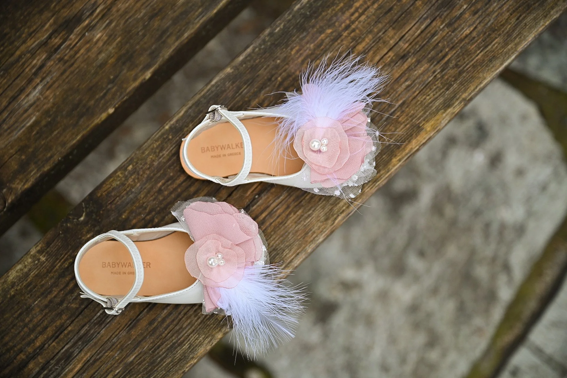 Pair of white baby shoes with pink fabric flowers, pearl decorations, and white feathers, placed on a wooden surface.