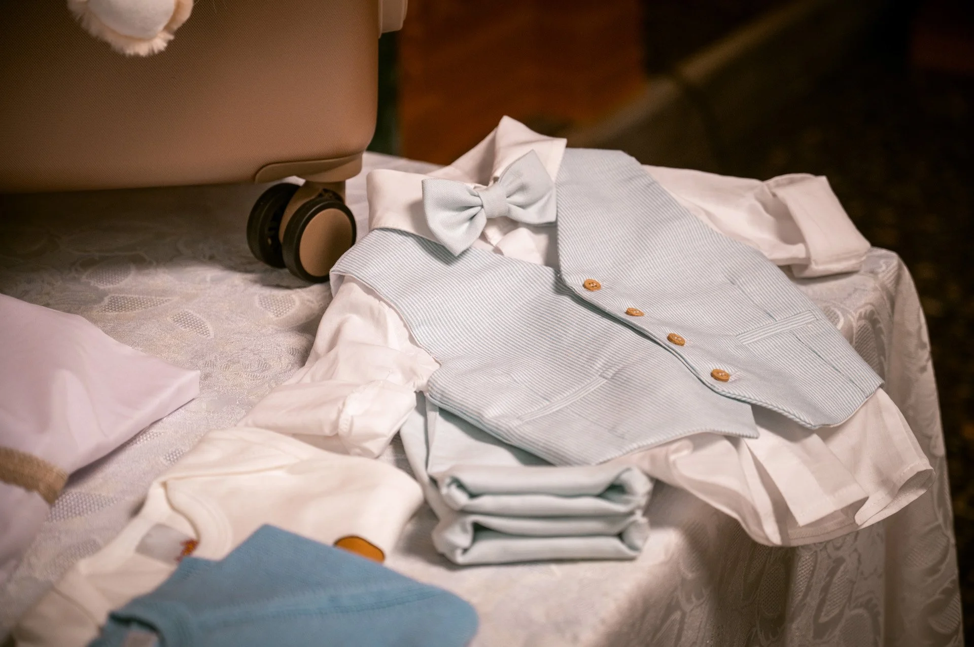 A collection of children's formal clothing including a white shirt, light blue vest, matching bow tie, and pants, arranged on a table with a suitcase in the background.