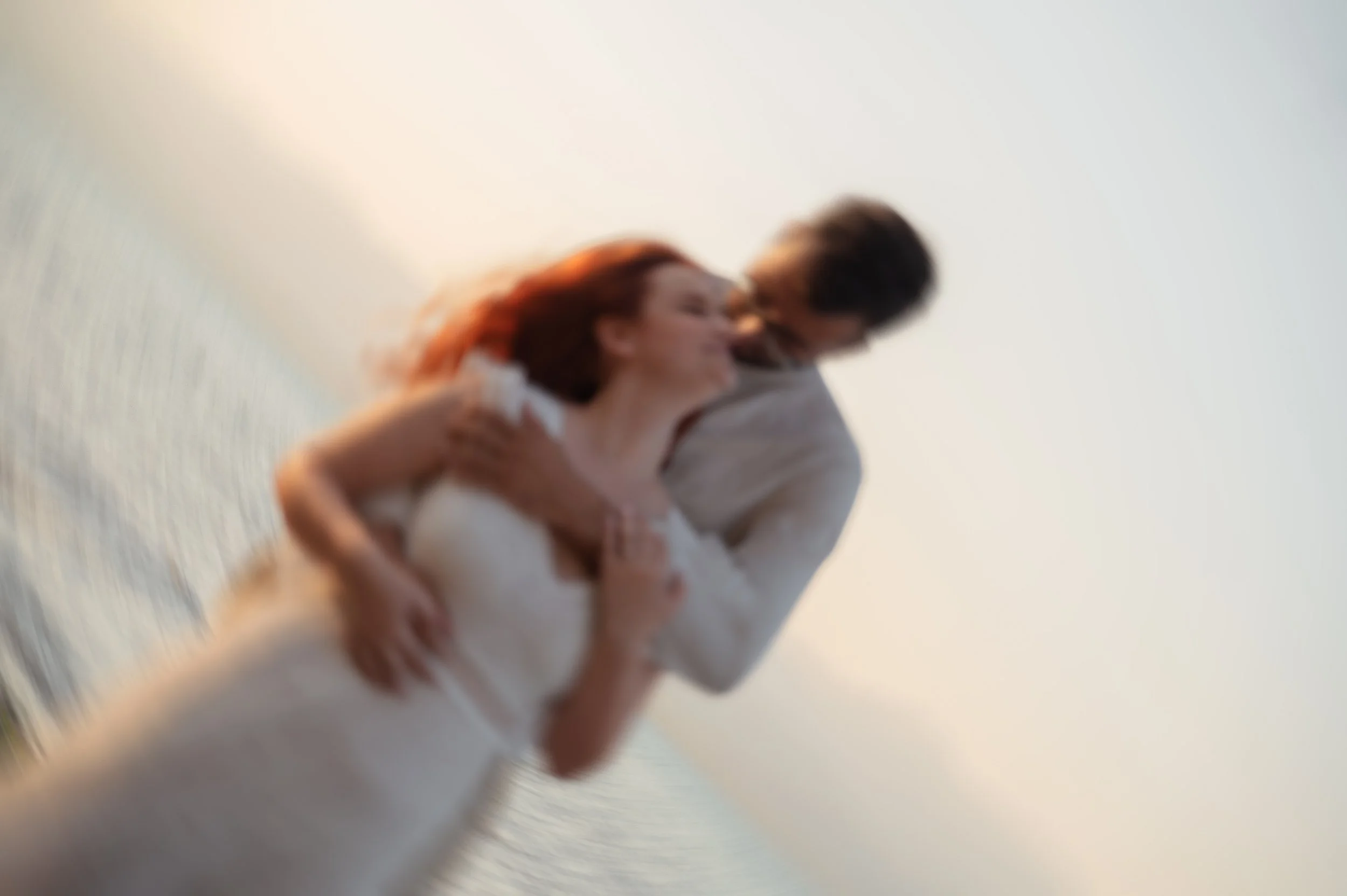 A blurry photo of a couple, with the woman wearing a white dress with lace details and the man in a light-colored shirt, embracing near water with a soft sky background.