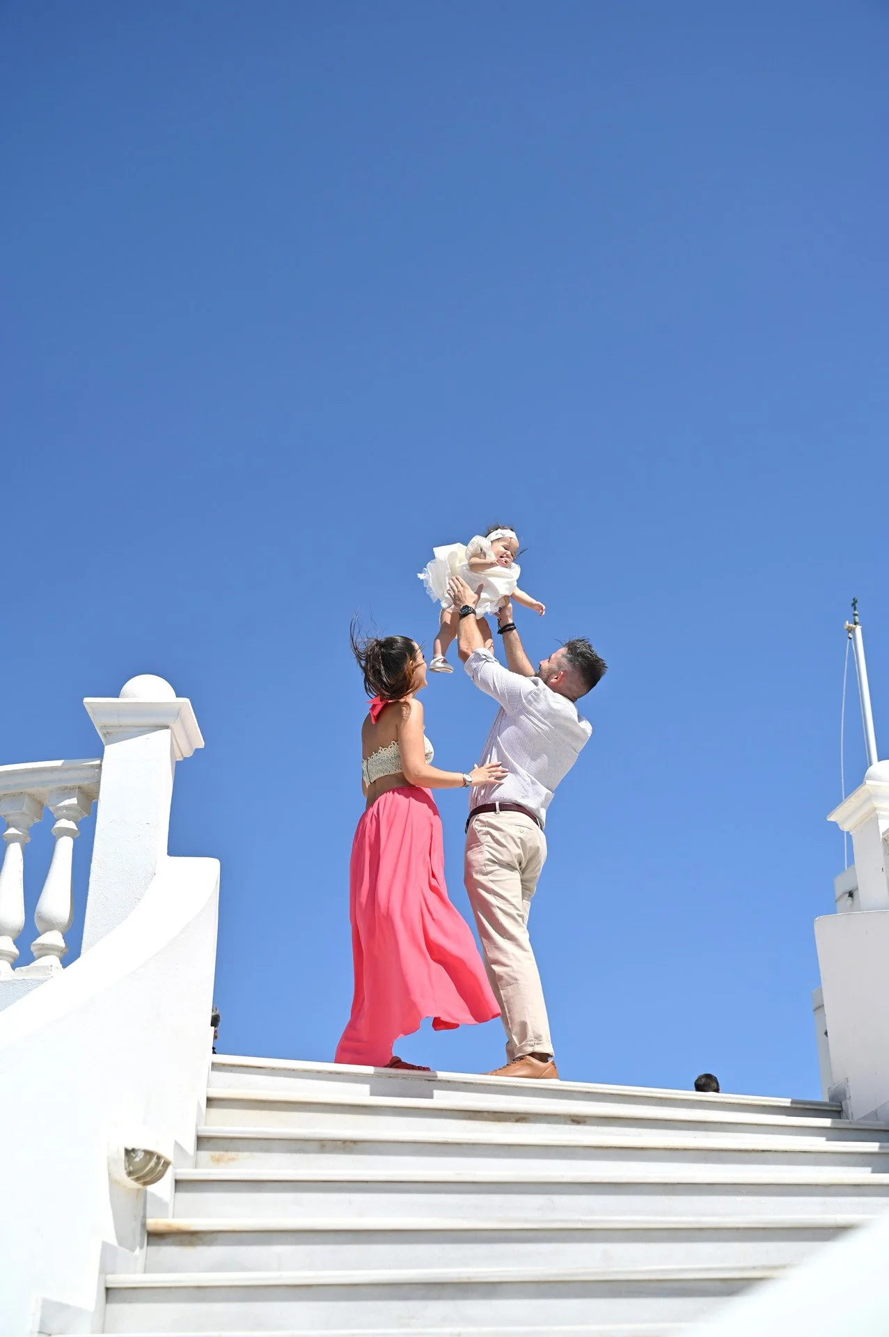 A man, woman, and young girl are on white stairs outdoors, with the man lifting the girl into the air against a clear blue sky.