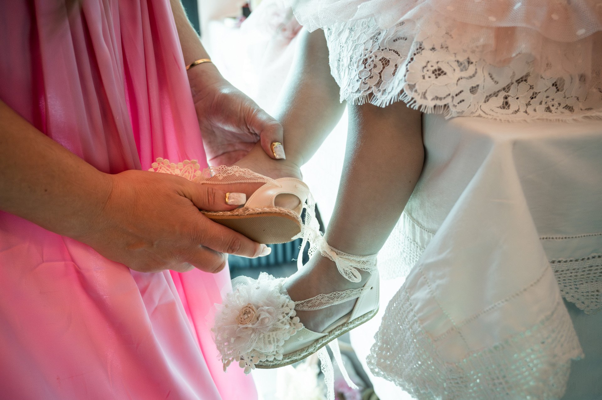A woman in a pink dress helping a young girl in a white dress put on a pair of white shoes with lace details and a decorative flower.