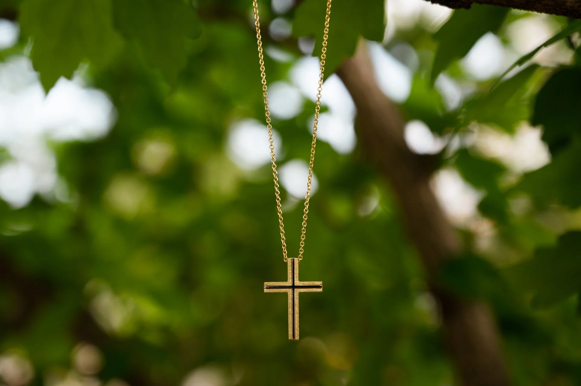 Gold cross necklace hanging from a tree branch with green leaves in the background.