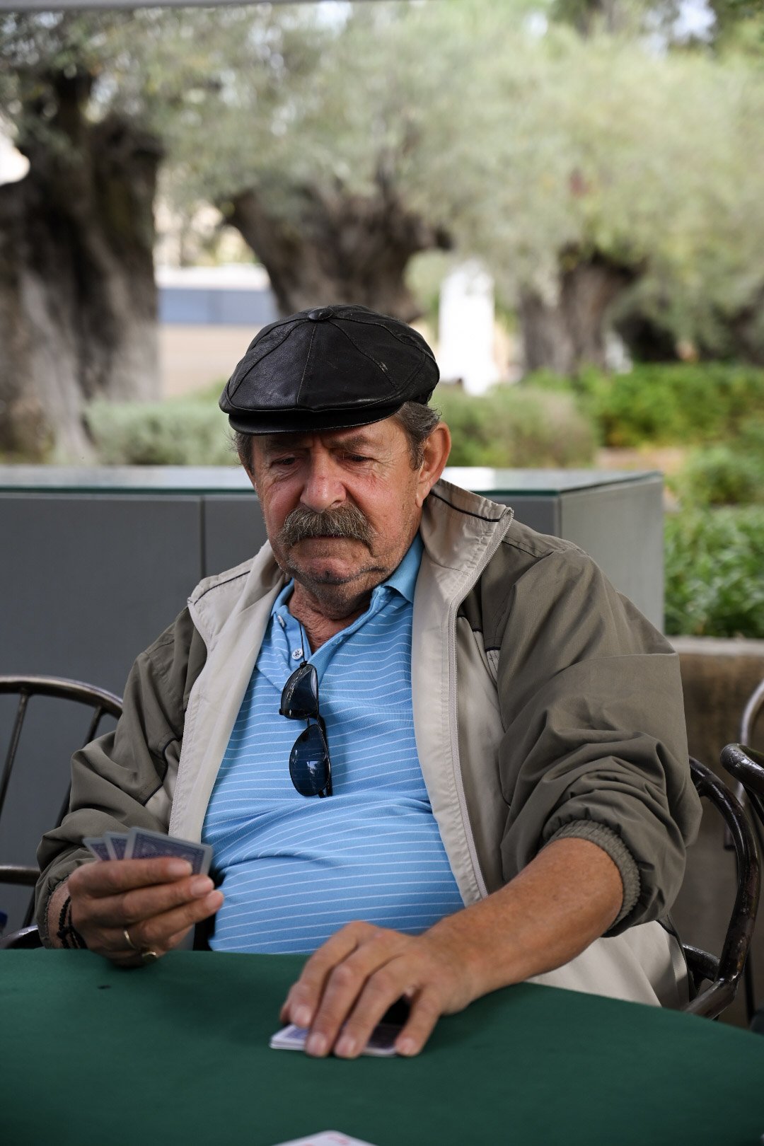 An older man with a mustache and sunglasses hanging on his shirt plays cards at a green table outdoors, wearing a gray jacket, a blue striped polo shirt, and a black cap.