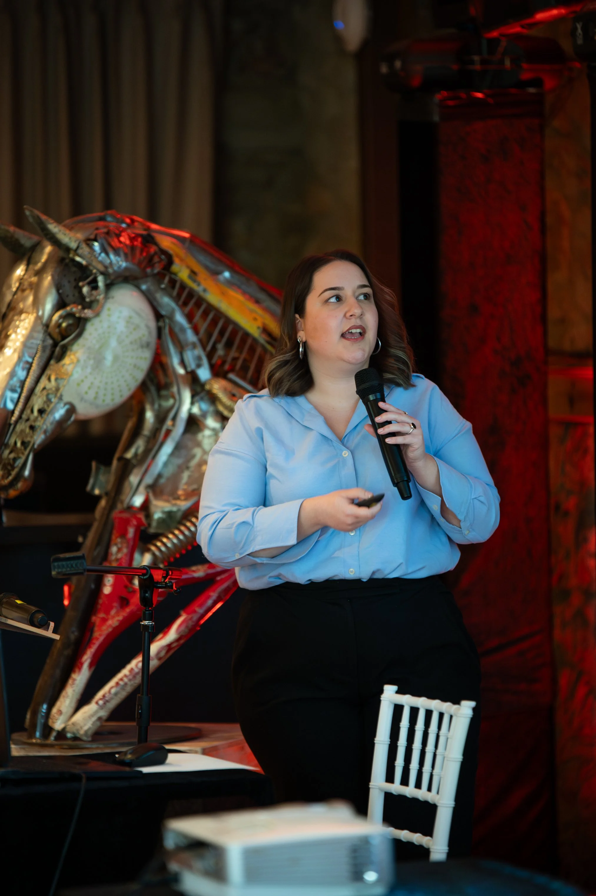 A woman in a blue shirt holding a microphone and speaking at an event, with a large decorative horse sculpture behind her.