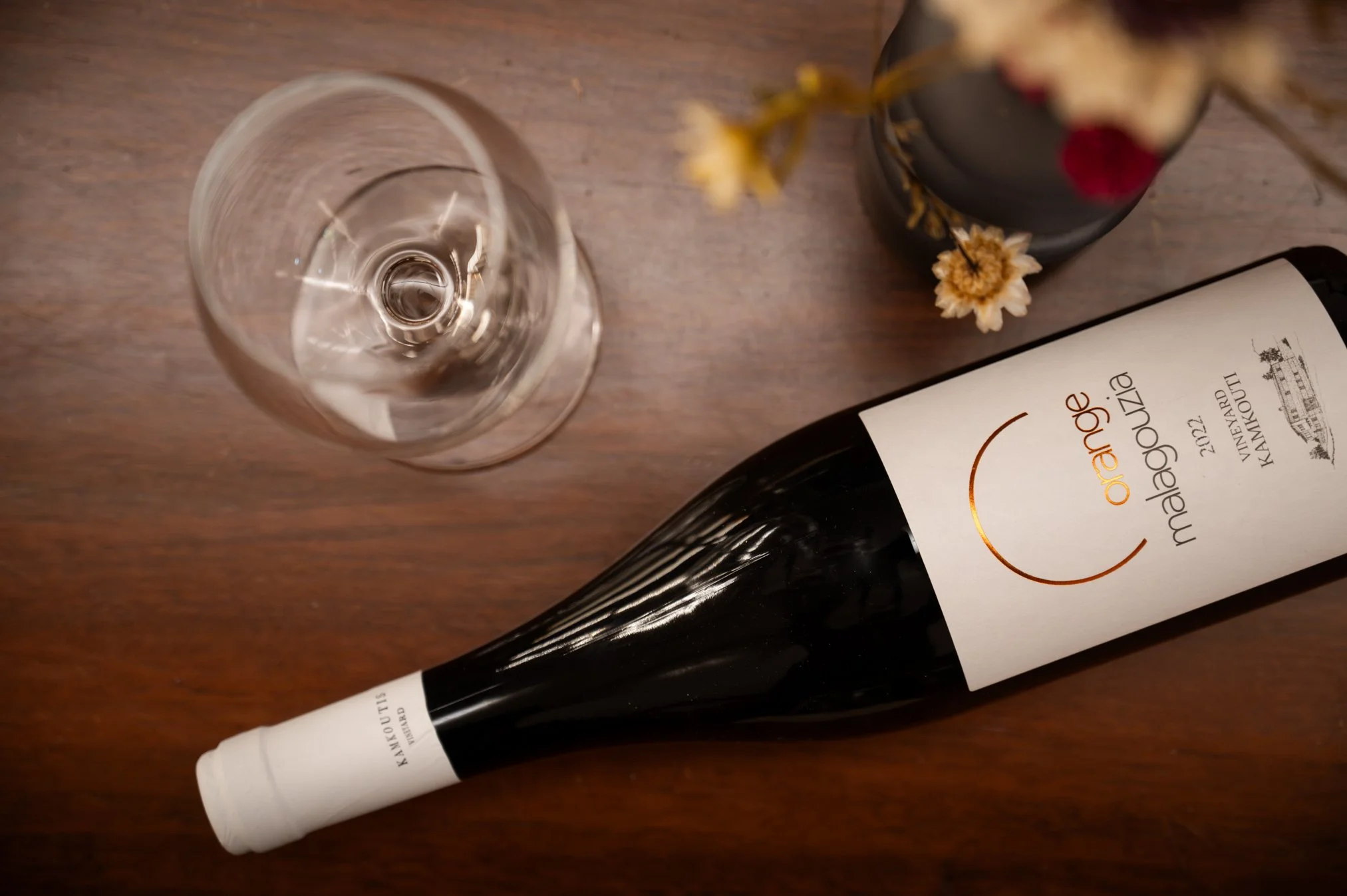 A bottle of orange malagousia wine lying on a wooden table with a wine glass next to it and a small vase with dried flowers.