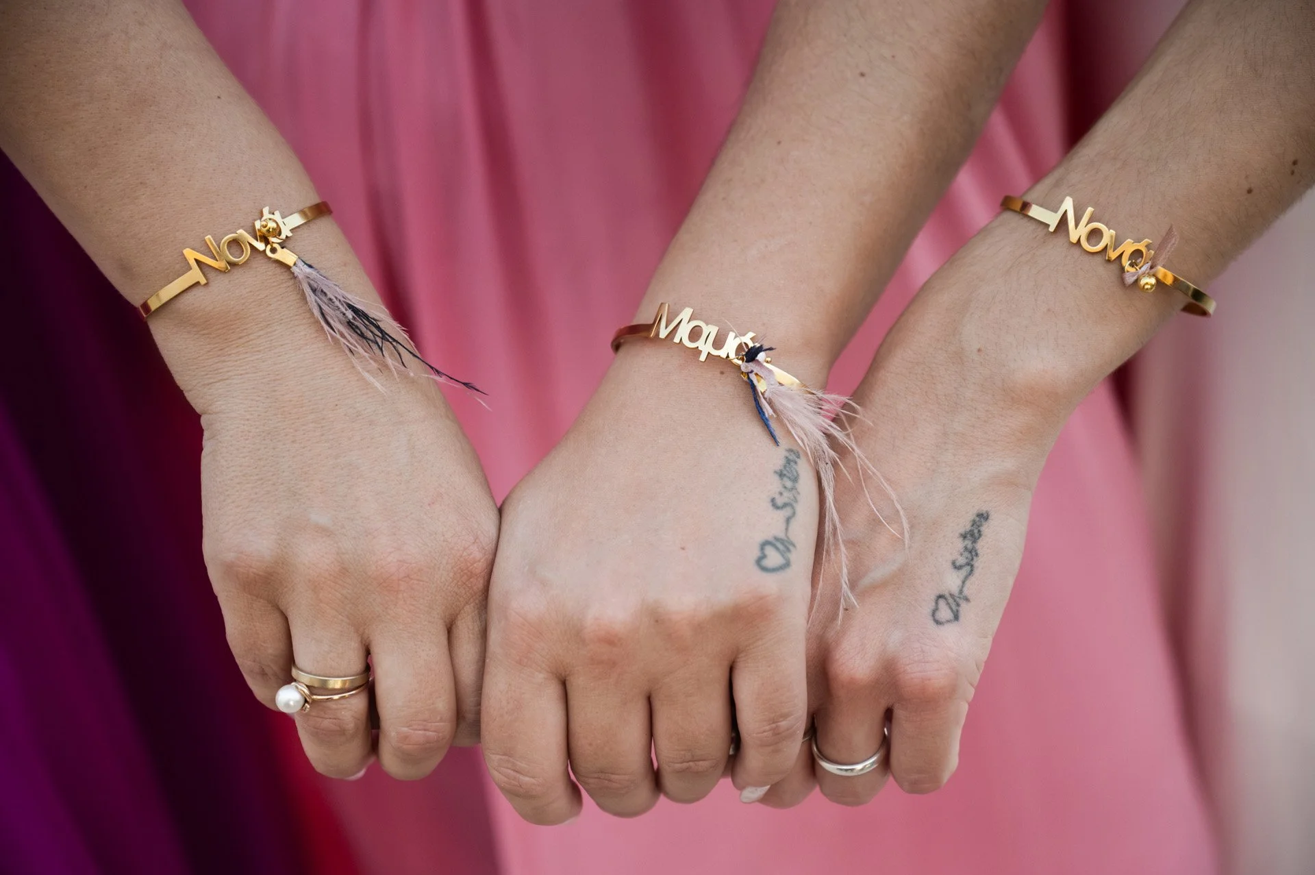 Three hands with matching gold bracelets that say 'Nora' and small feather charms. The hands are linked, showing rings and tattoos.