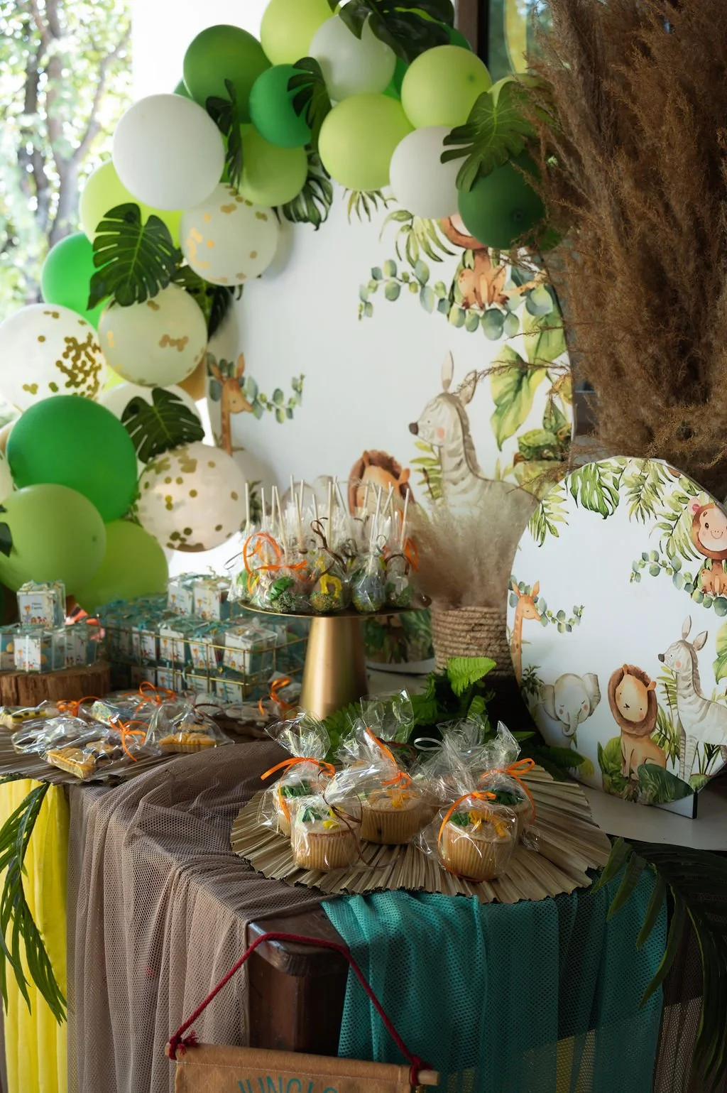 Decorations for a jungle or safari-themed party with balloons, cutouts of jungle animals, wrapped cupcakes, and party favors on a table.