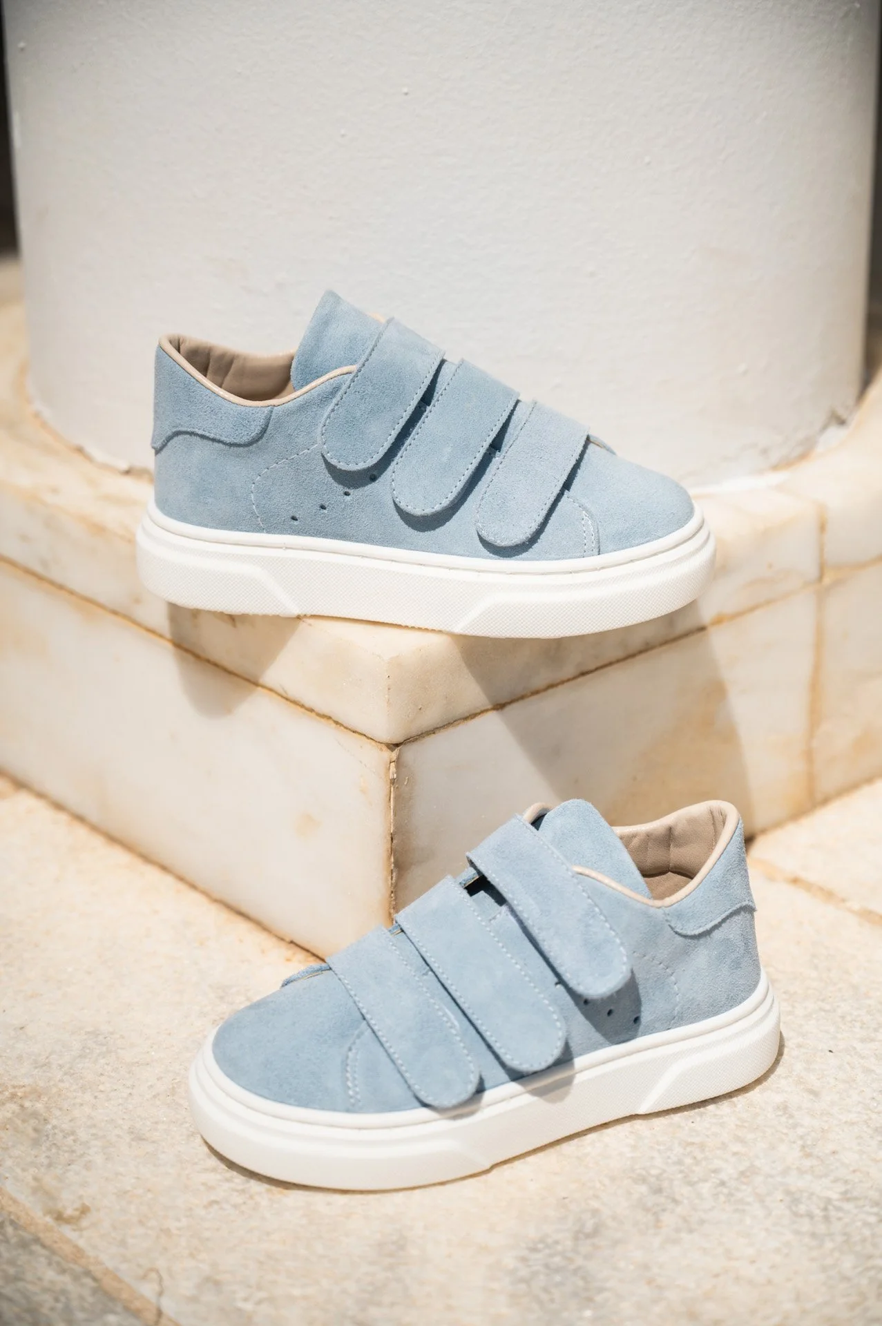 Pair of light blue suede sneakers with velcro straps placed on a stone surface and a marble block in the background.