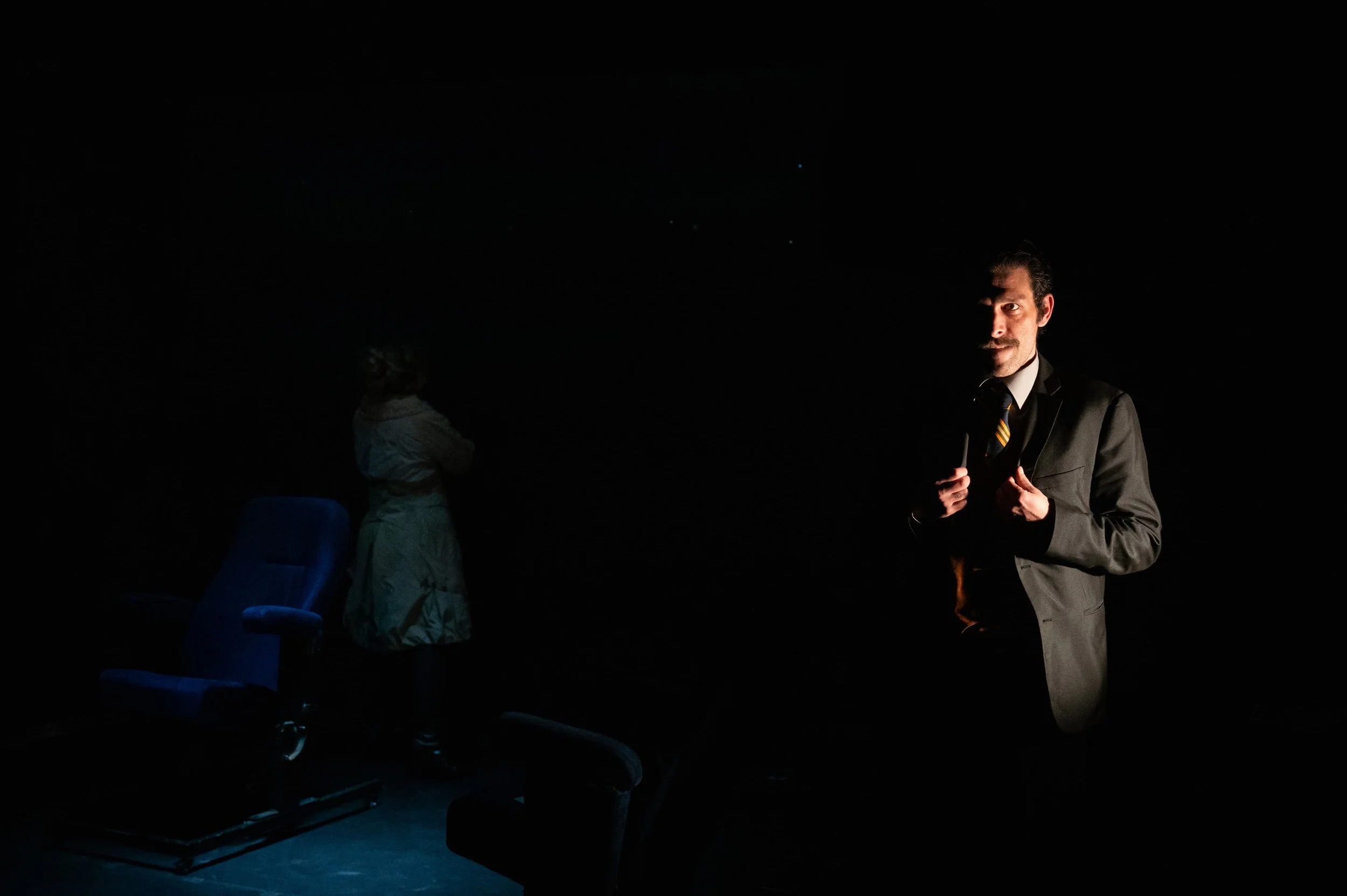 A man in a suit with a tie stands in a dark room, illuminated by a single light source, with a woman in the background facing away from the camera and a blue office chair nearby.