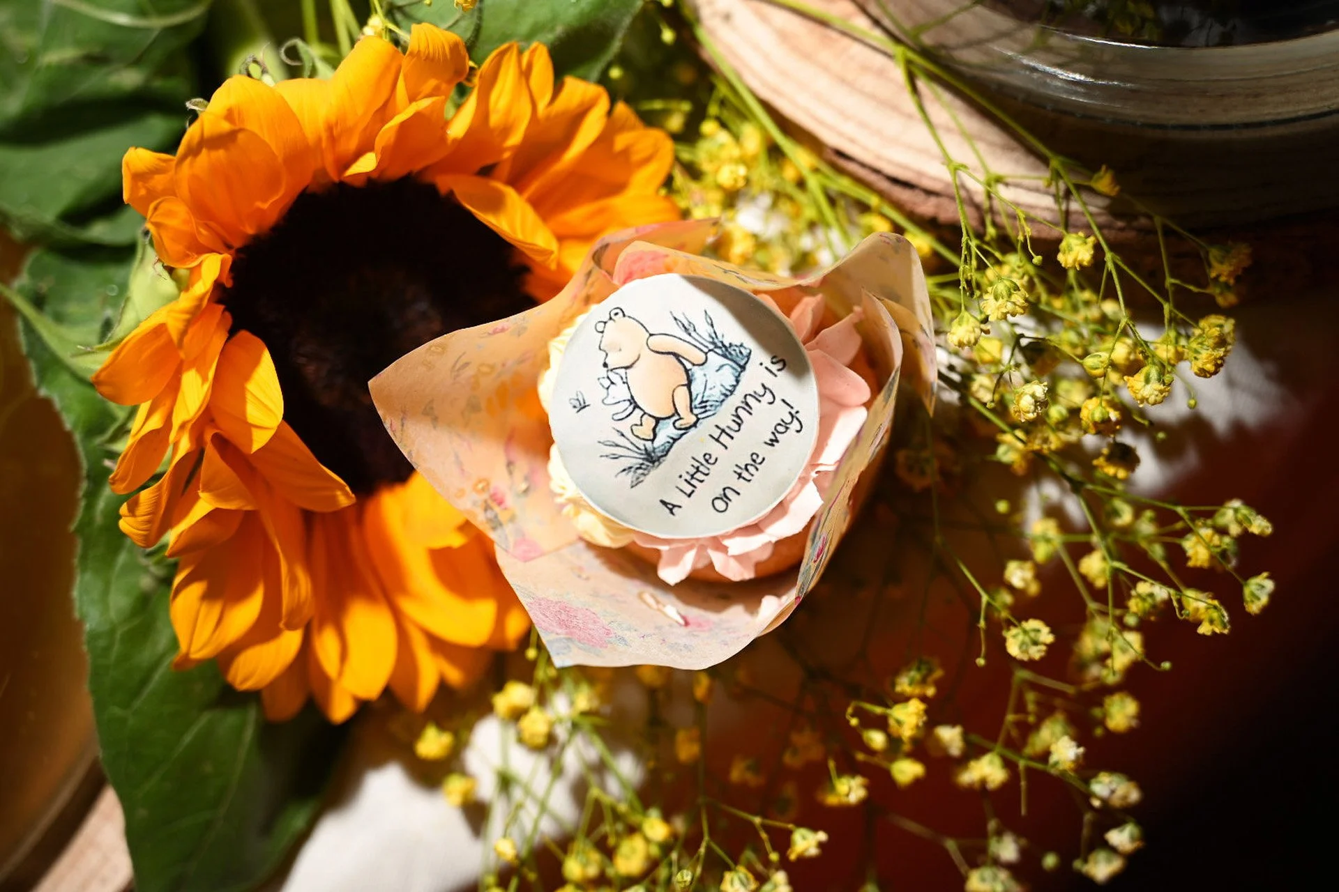 A sunflower with bright yellow petals and dark center next to a small bouquet wrapped in tissue paper with a Winnie the Pooh sticker that reads 'A Little Honey is On the way!' surrounded by small yellow flowers.