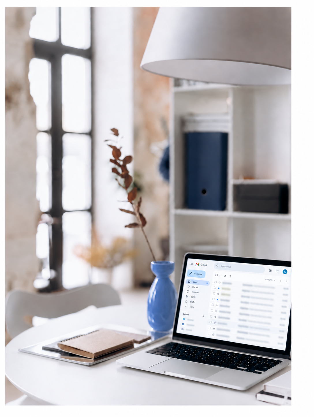 A white desk with an open laptop displaying an inbox email interface, a blue vase with a dried brown leaf, and notebooks, in a bright room with a large window and a white shelving unit in the background.