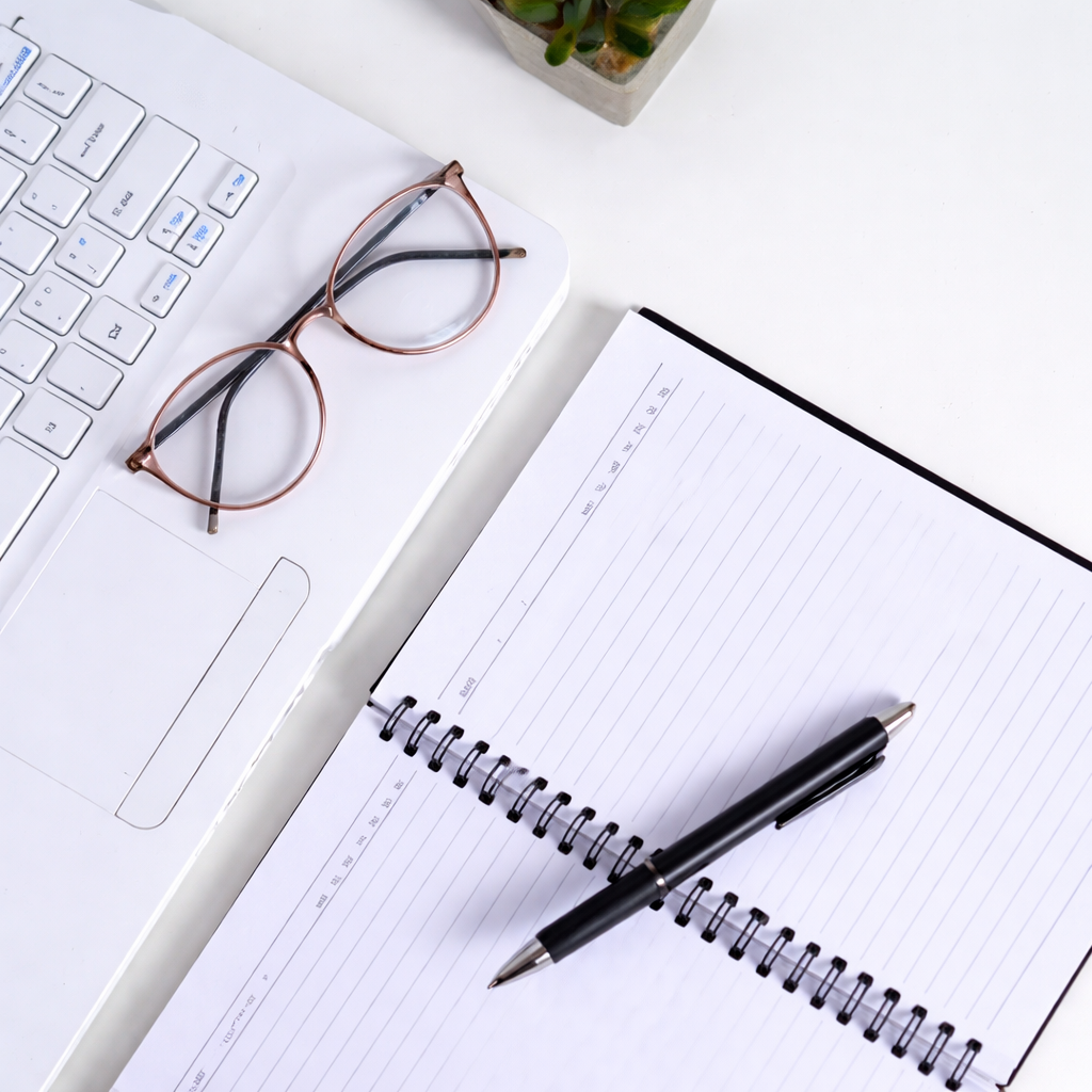 A workspace with a white keyboard, a pair of glasses, a black pen, an open lined notebook, and a small potted plant on a white surface.