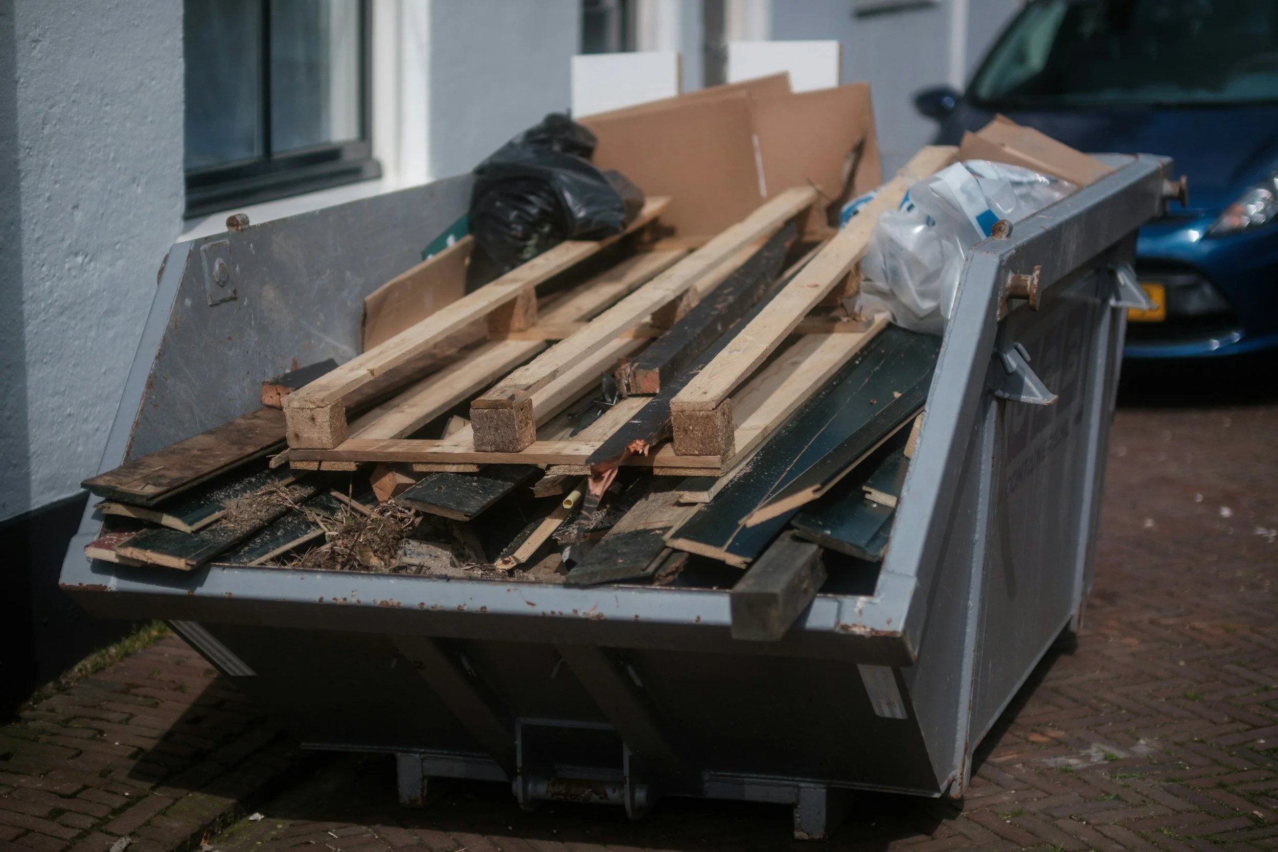 A large open dumpster filled with used wood planks, cardboard, and trash bags outdoors, next to a brick sidewalk and building wall, with a blue car partially visible in the background.