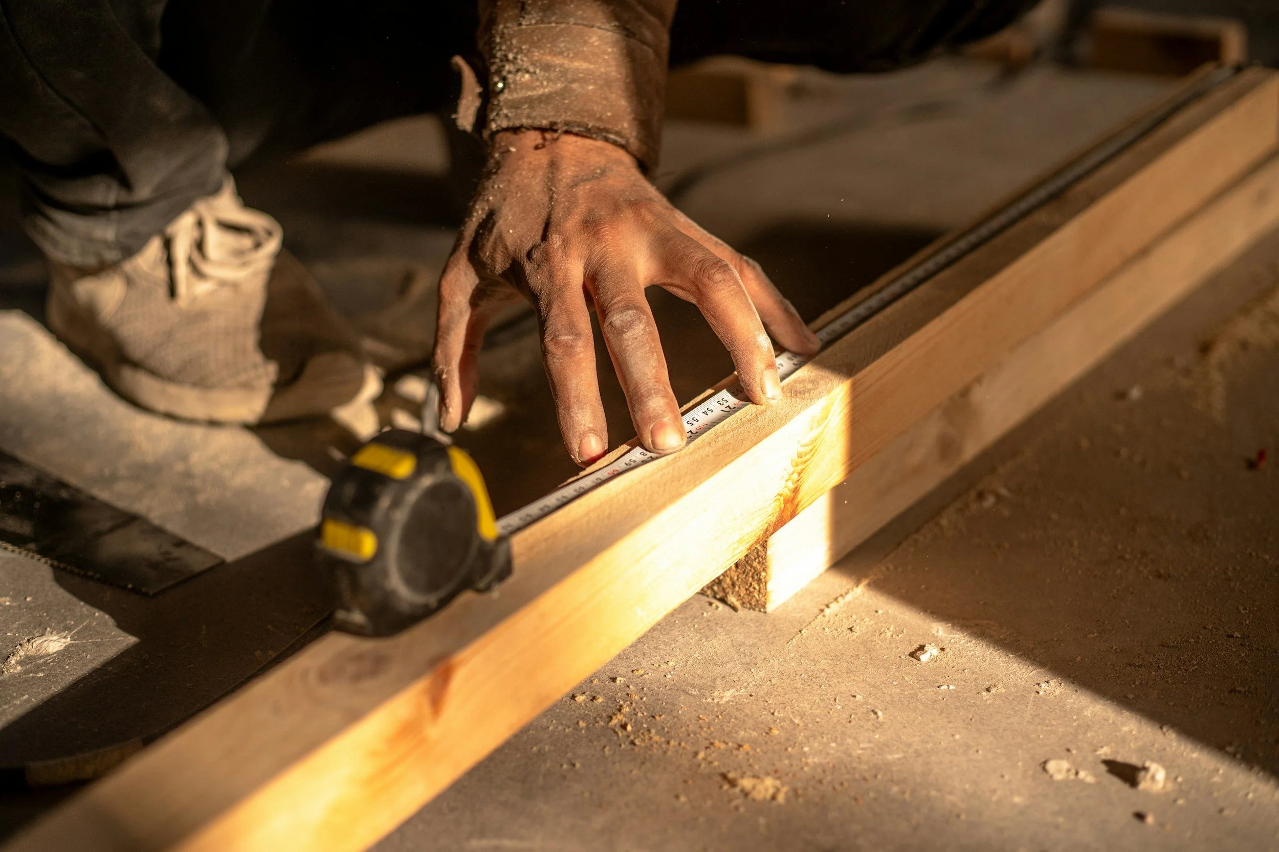 Person measuring a piece of wood with a tape measure, bending down on a construction site.