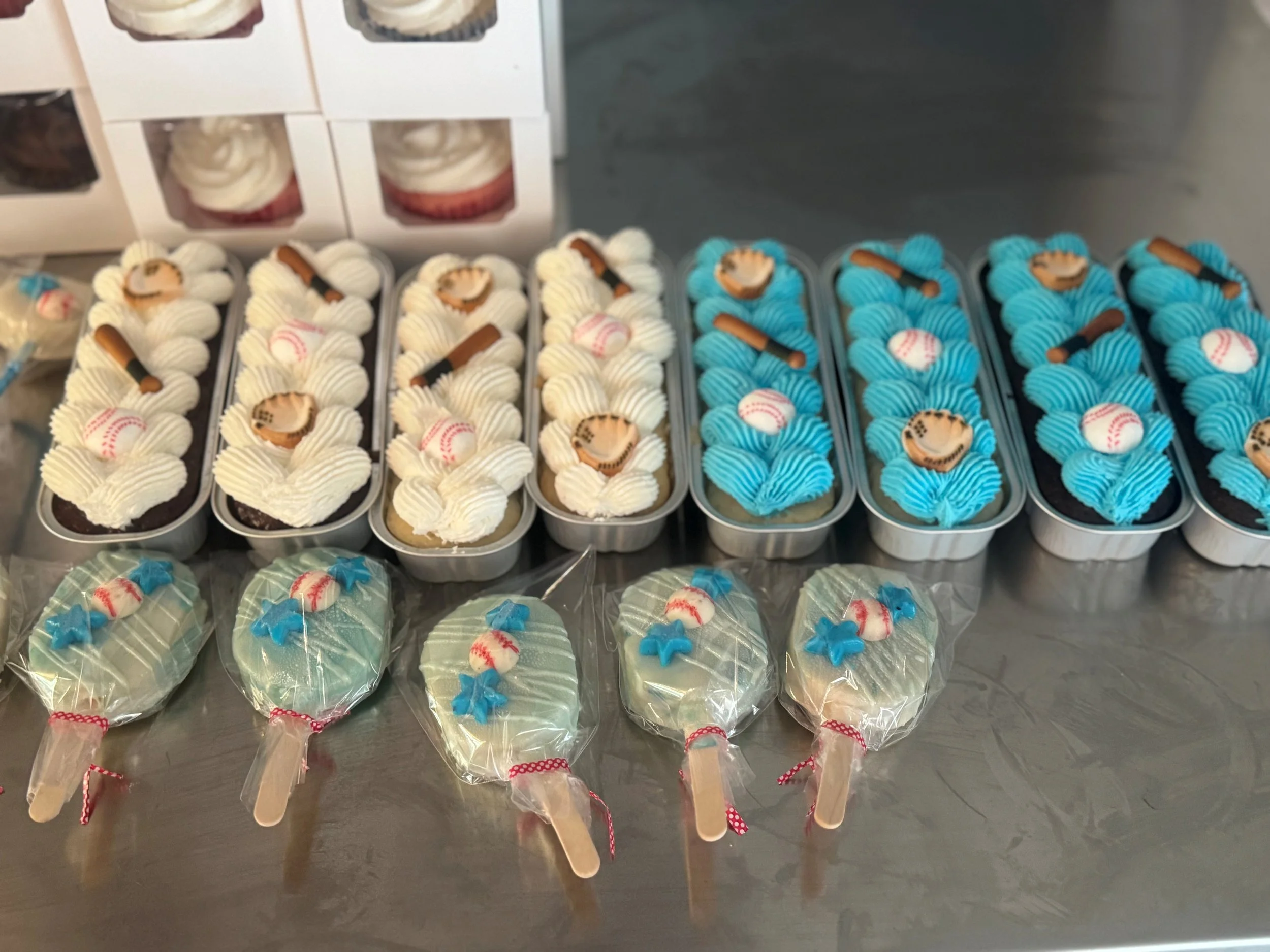 Multiple baseball-themed ice cream pops with blue or white icing, some decorated with small baseball ornaments and colored ribbons, displayed on a table.