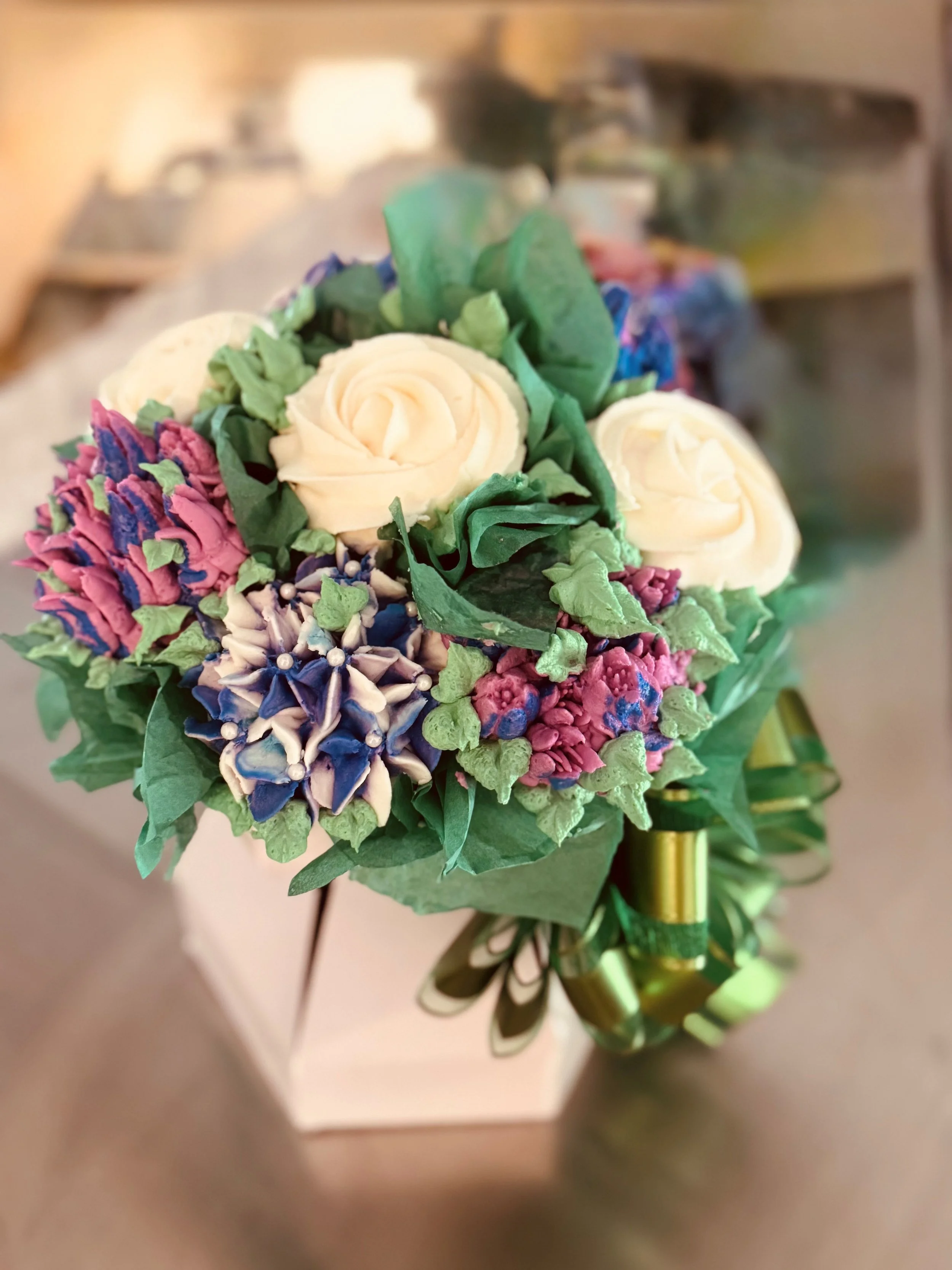 A bouquet of artificial flowers including cream roses, purple and pink hydrangeas, and greenery, wrapped in green paper and decorated with pearl beads.