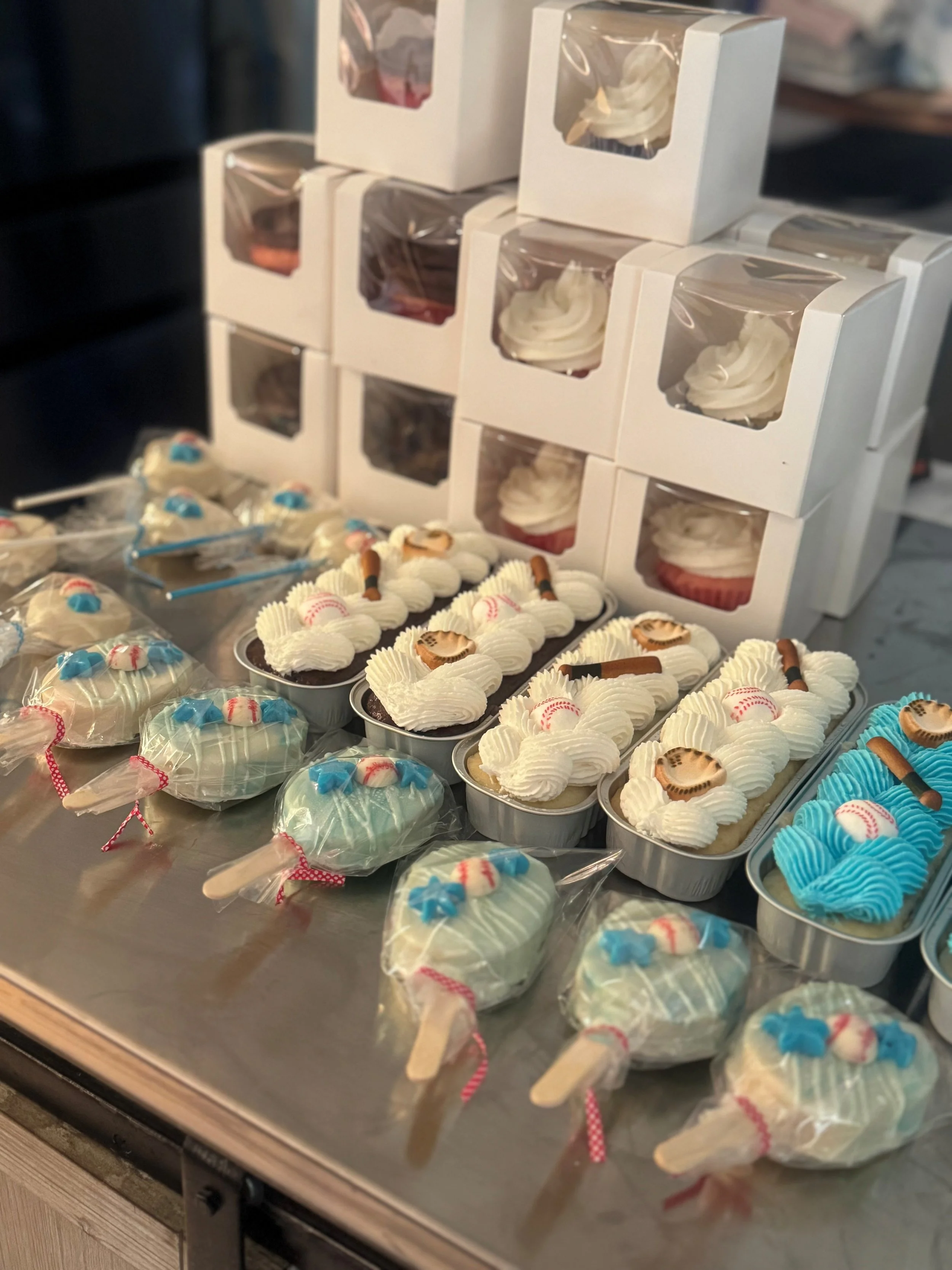 Various cupcakes on display, decorated with white, blue, and pink icing, some wrapped in plastic, with boxed cupcakes stacked in the background.