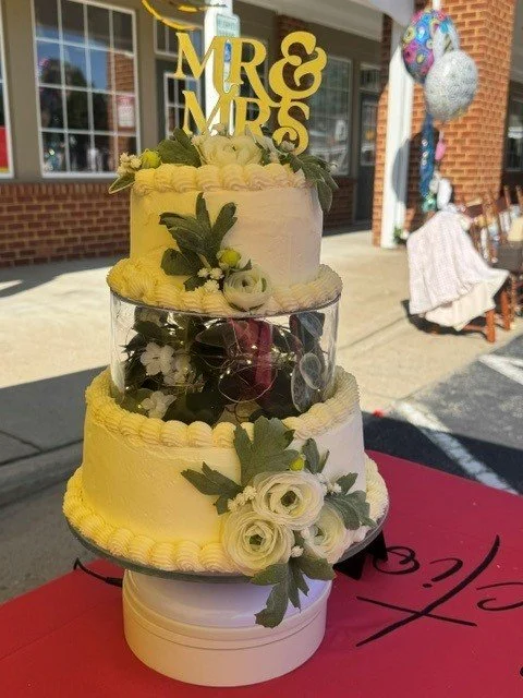 Three-tier wedding cake with white and yellow frosting, decorated with white roses and green leaves, topped with gold 'Mr & Mrs' cake topper, placed on a table outside. In the background, there are balloons and a person sitting at a table.