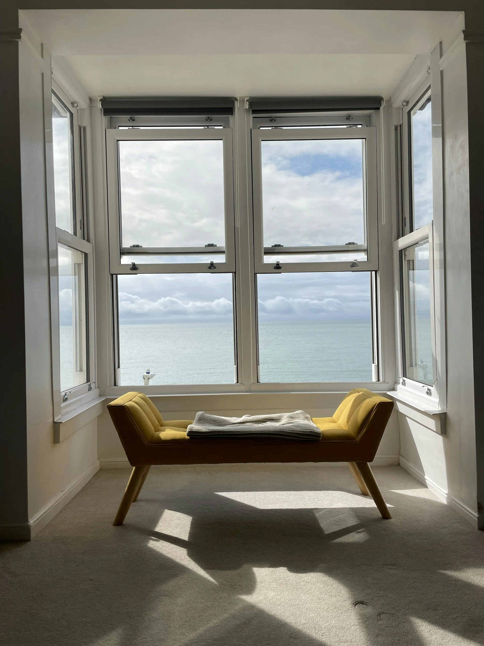 A room with large windows overlooking the ocean, with a yellow upholstered bench and a folded blanket on top, and cloudy sky visible outside.