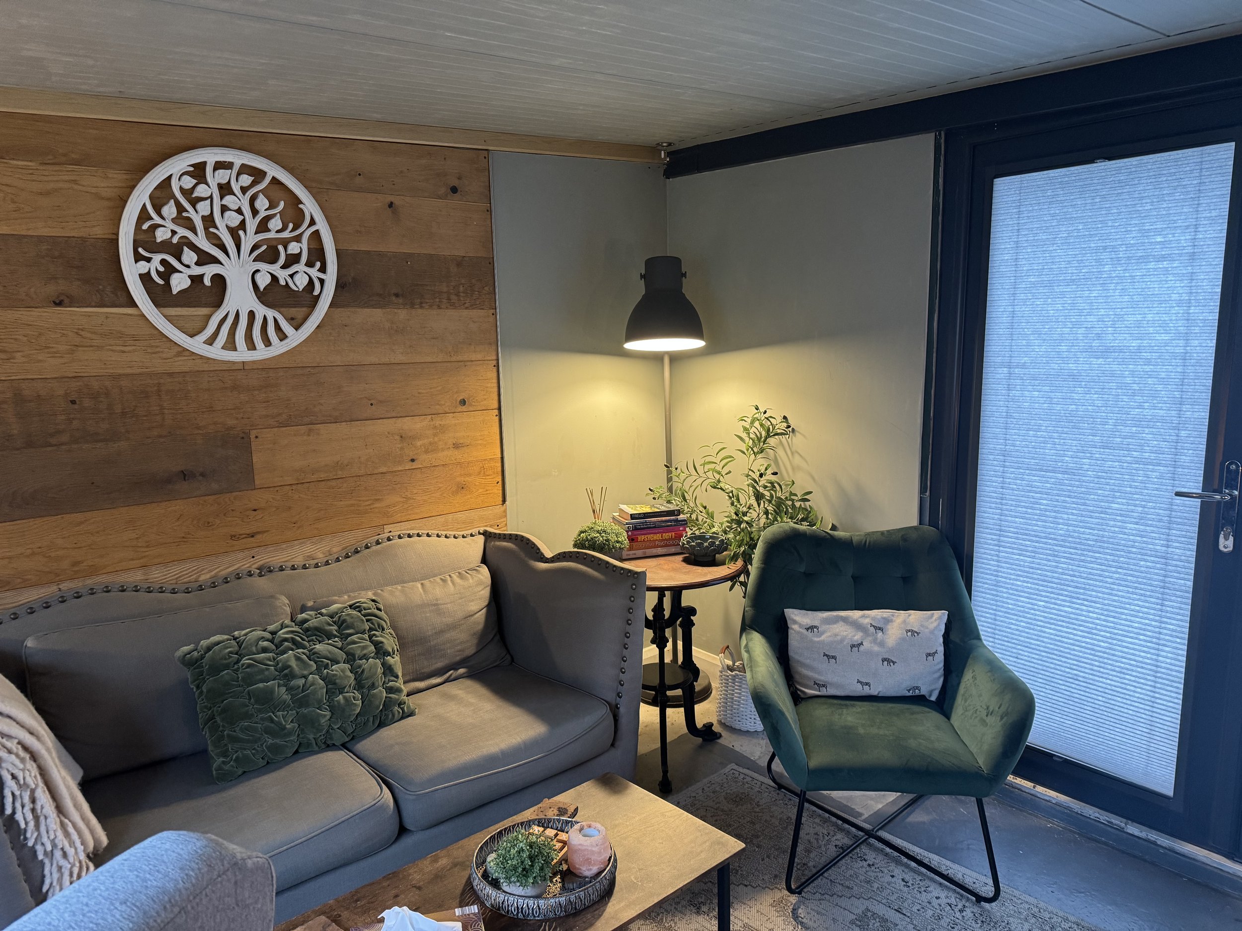 A cozy living room corner with a beige couch, a green velvet armchair with a patterned pillow, a wooden side table with books and plants, a black floor lamp, a decorative wall art of a tree, and a large door with a screen on it.