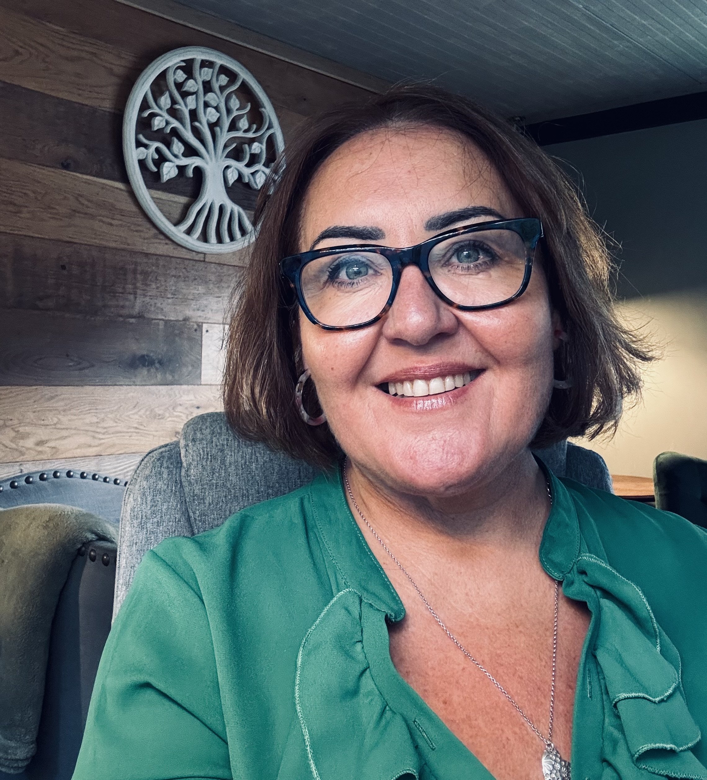 Smiling woman with short brown hair, glasses, and green blouse, sitting in a room with wooden wall decor and a cozy chair.