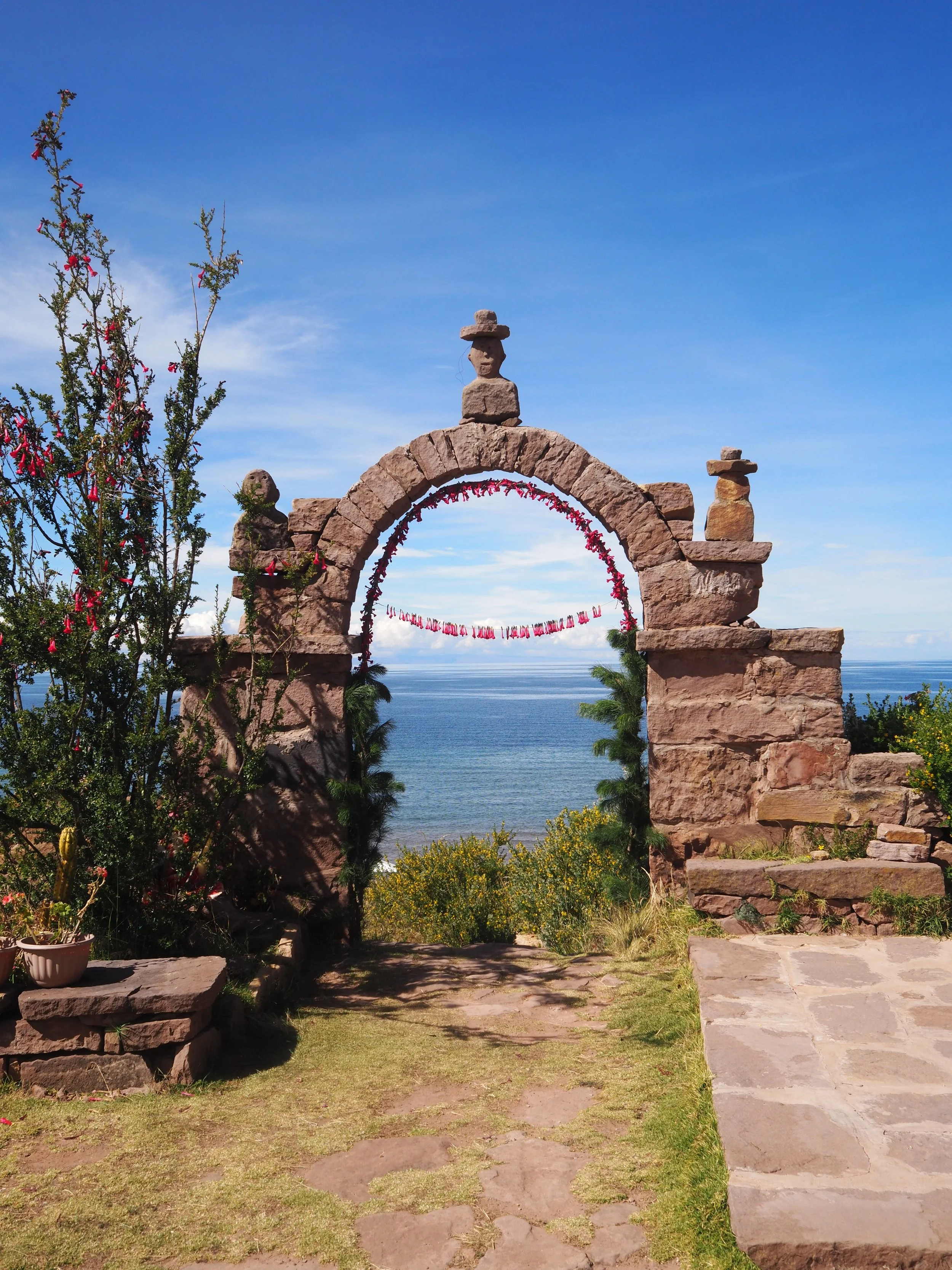 Stone archway overlooking a body of water with a clear blue sky in the background. The arch is decorated with pink ribbons, and there are plants and flowers around it.