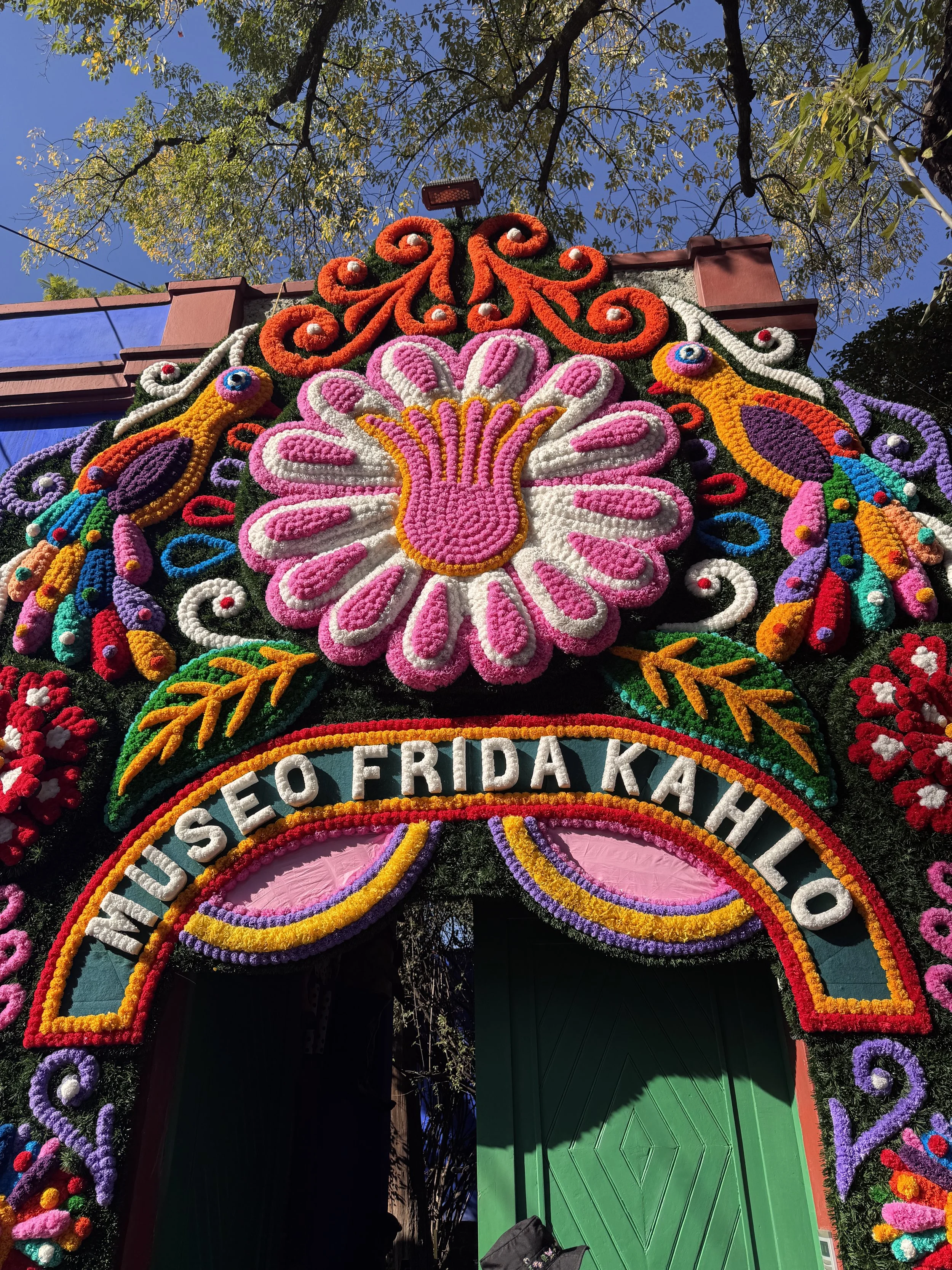 Colorful floral tribute with the words "NIUO E O FRIDA KAHLO" at the entrance of a decorative arch, featuring a large pink and white flower, peacocks, and vibrant patterns, set against a blue sky and green trees. Frida Kahlo Museum