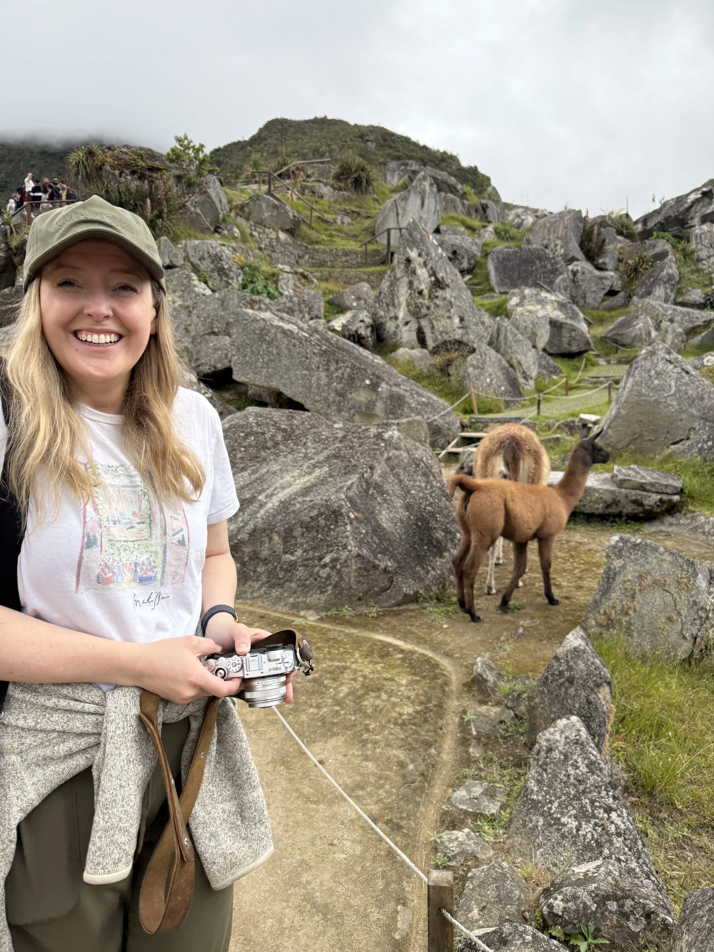 A woman smiling and holding a camera on a trail with rocky terrain, alpacas grazing nearby, and a group of people in the background.