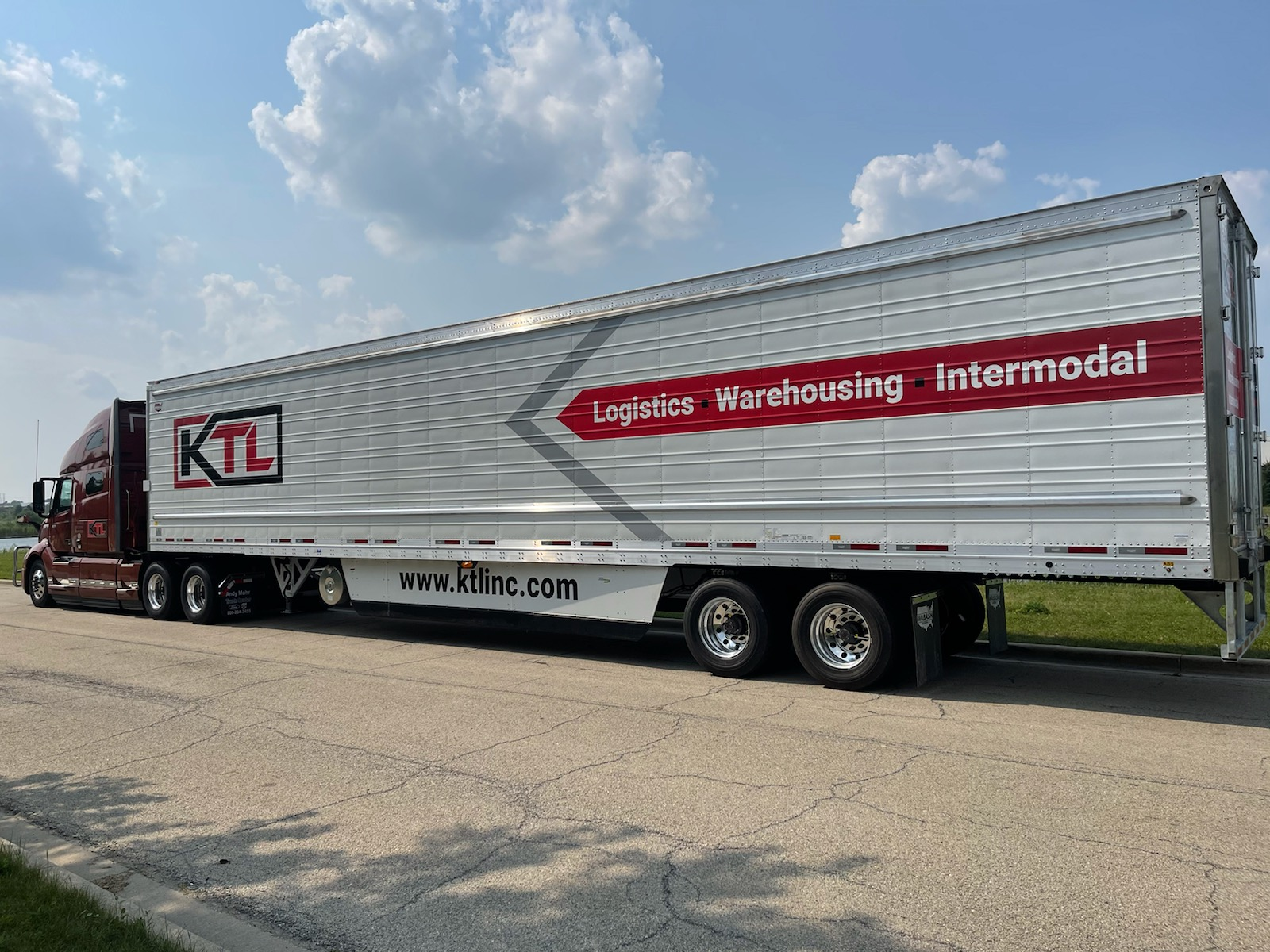 A large semi-truck with a red cab and a white trailer parked on a paved road under a partly cloudy sky. The trailer has the logo 'KTL' and text indicating logistics, warehousing, and intermodal services, along with a website URL.